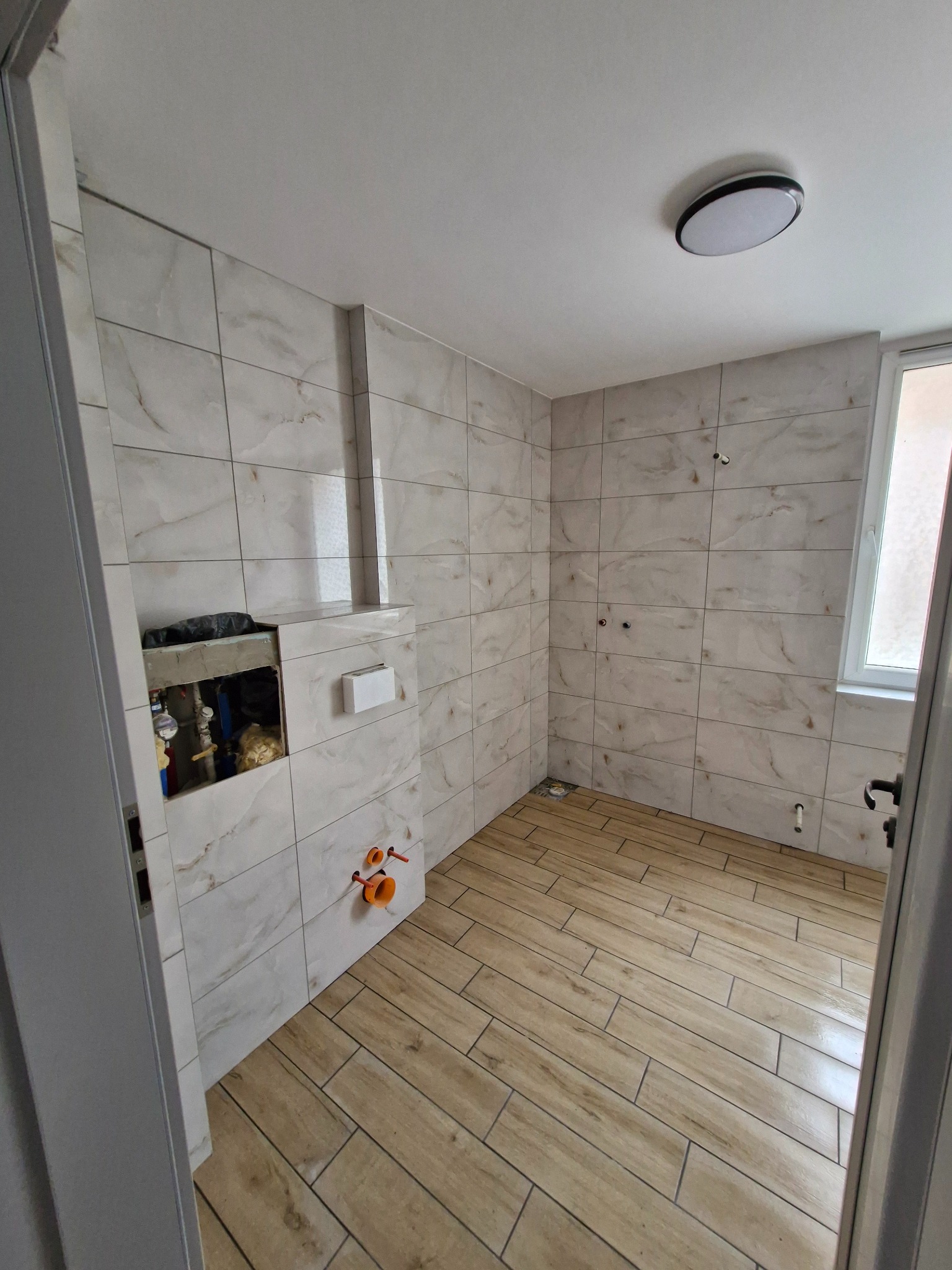 Wnętrze łazienki w trakcie prac: ułożone płytki imitujące drewno na podłodze, częściowo wykończone ściany w jasnych płytkach. Widoczne instalacje i otwór w ścianie.