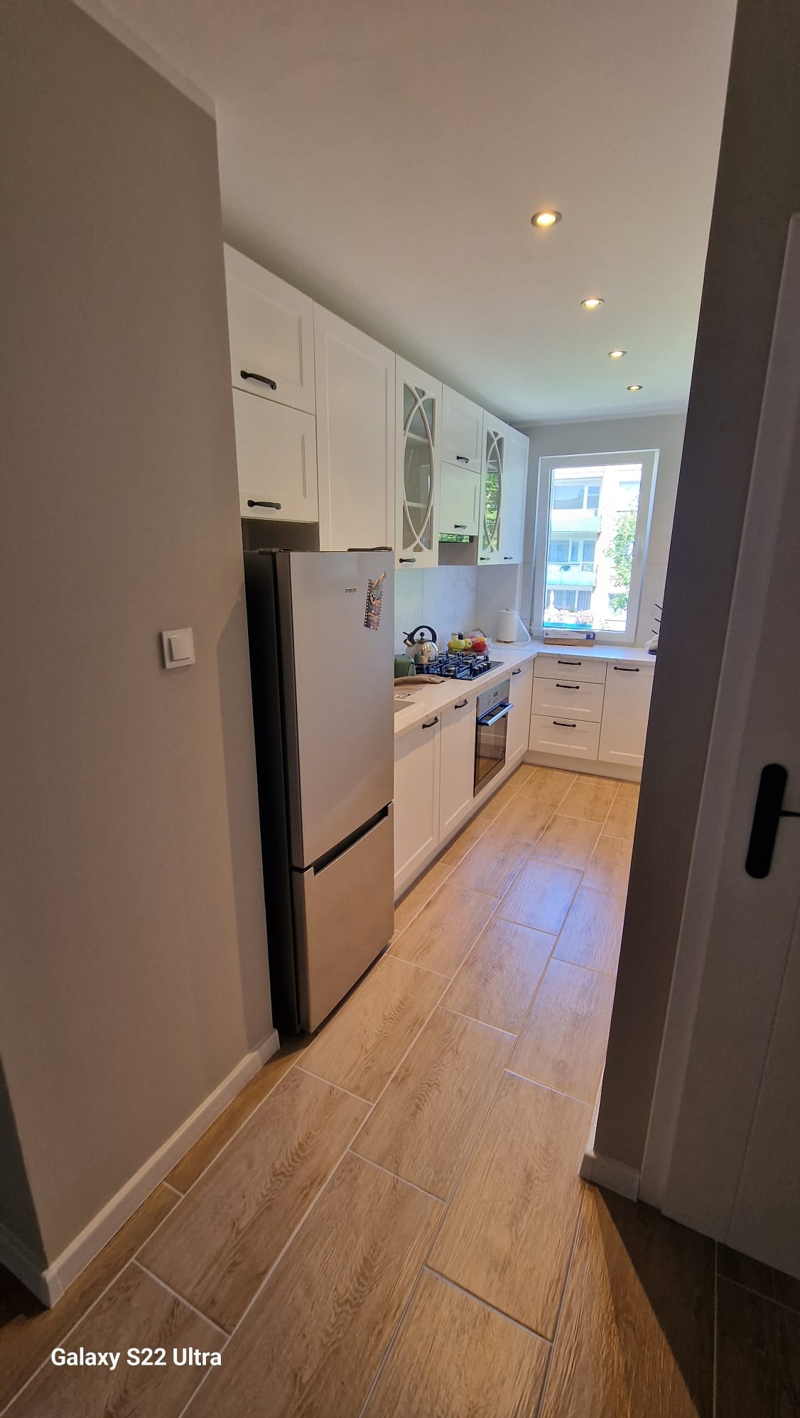 Nowoczesna kuchnia z białymi szafkami, srebrną lodówką i drewnopodobnymi płytkami podłogowymi. Widok z perspektywy wejścia, oświetlenie sufitowe.