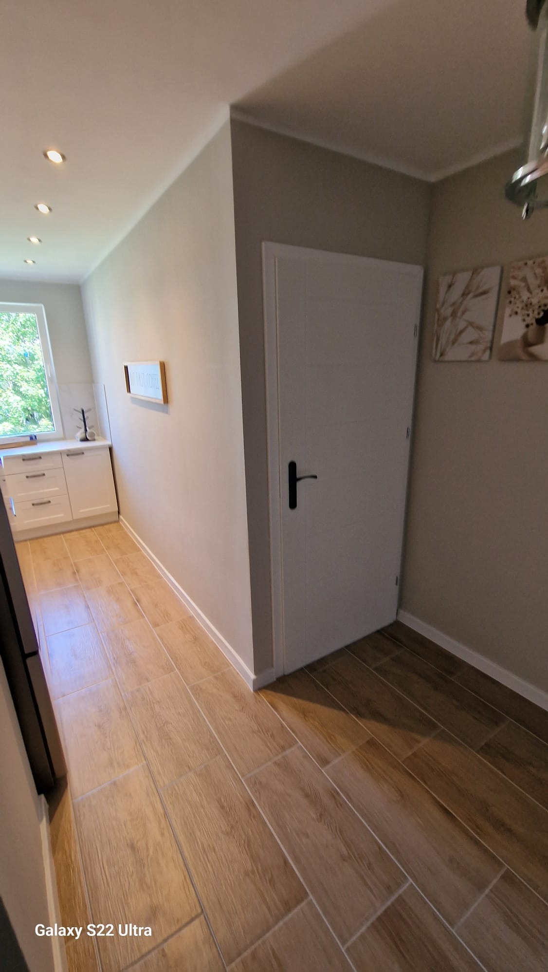 Jasne płytki podłogowe imitujące drewno w korytarzu, białe drzwi z czarną klamką, ściany w odcieniach beżu i szarości, dekoracje ścienne.