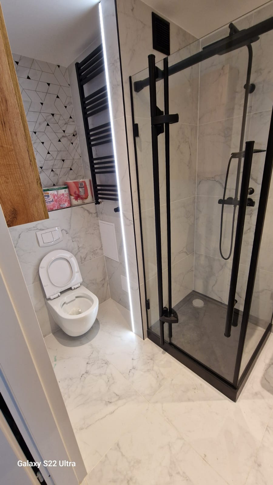 Wykończona łazienka z kabiną prysznicową w czarnej ramie, toaletą i grzejnikiem drabinkowym. Białe płytki imitujące marmur i oświetlenie LED.