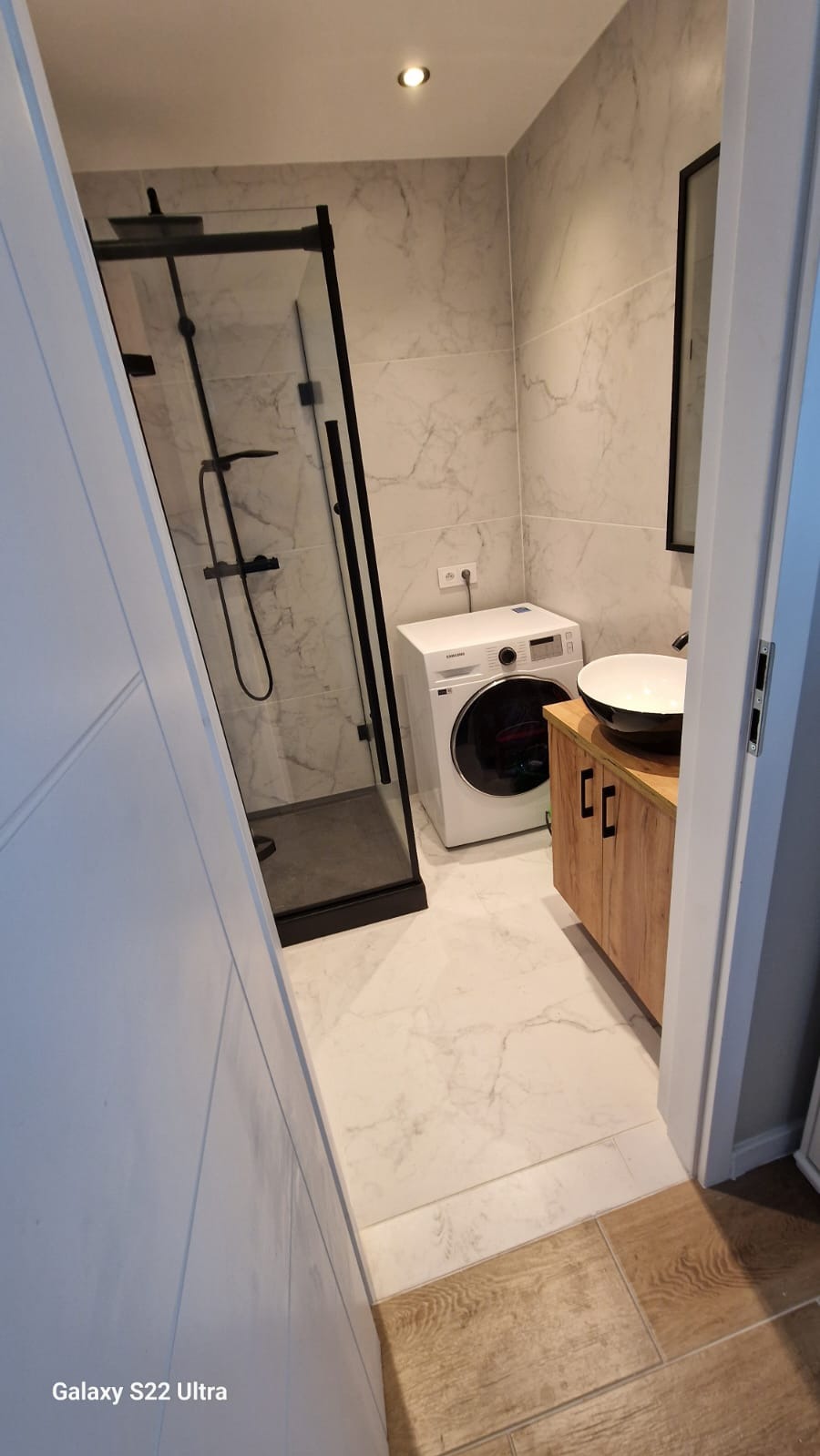 Nowoczesna łazienka z kabiną prysznicową typu walk-in, pralką i umywalką nablatową na szafce z drewnianymi frontami. Marmurowe płytki na ścianach i podłodze.