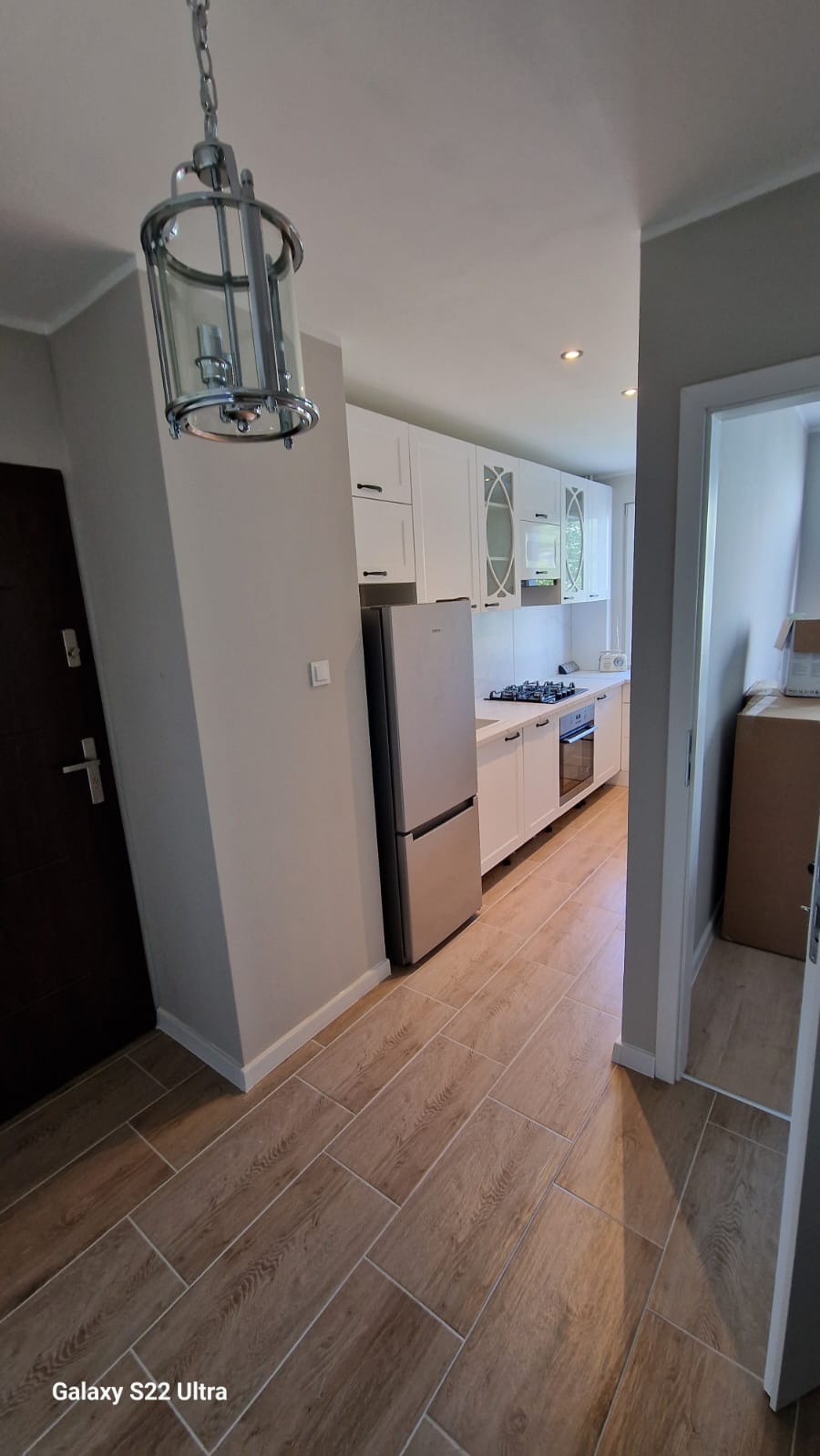 Nowoczesna kuchnia z białymi szafkami, srebrną lodówką i drewnopodobną podłogą z płytek, widziana z perspektywy wejścia. W tle widoczne okno.