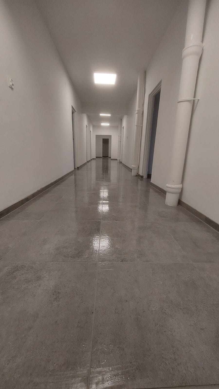 Długi, jasny korytarz z szarymi płytkami podłogowymi i białymi ścianami. Widoczne pionowe rury, drzwi i kwadratowe lampy sufitowe.