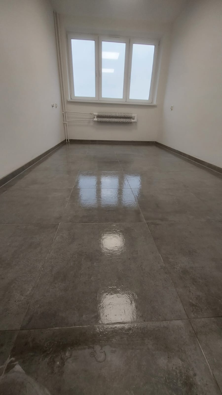 Pokój po remoncie z szarymi płytkami na podłodze, białe ściany i okno z grzejnikiem. Widok na gładką posadzkę z odbiciami światła okiennego.