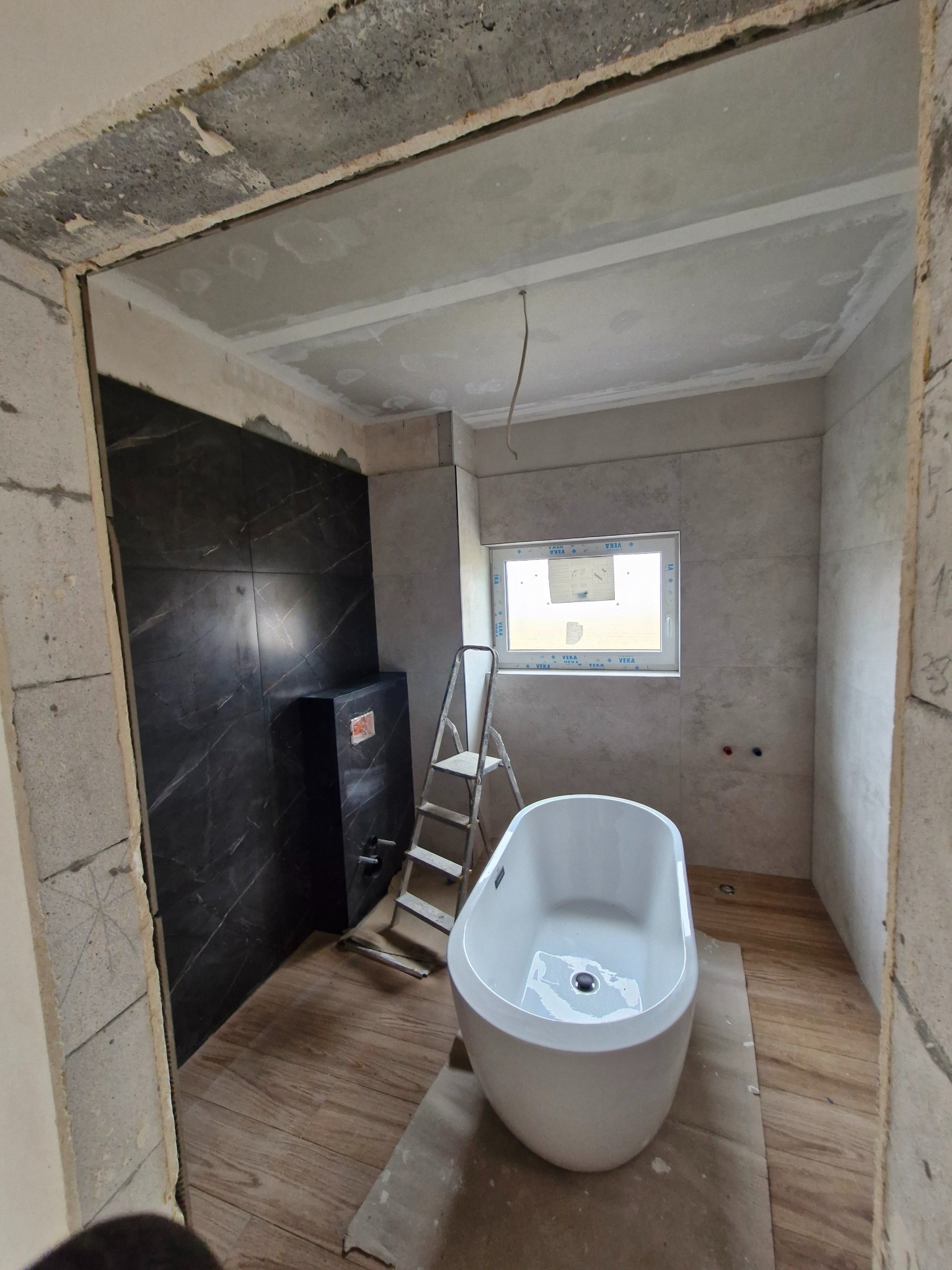 Wnętrze łazienki w trakcie prac: biała wanna wolnostojąca, czarne płytki imitujące marmur, drabina, okno. Widoczne niedokończone ściany z bloczków.