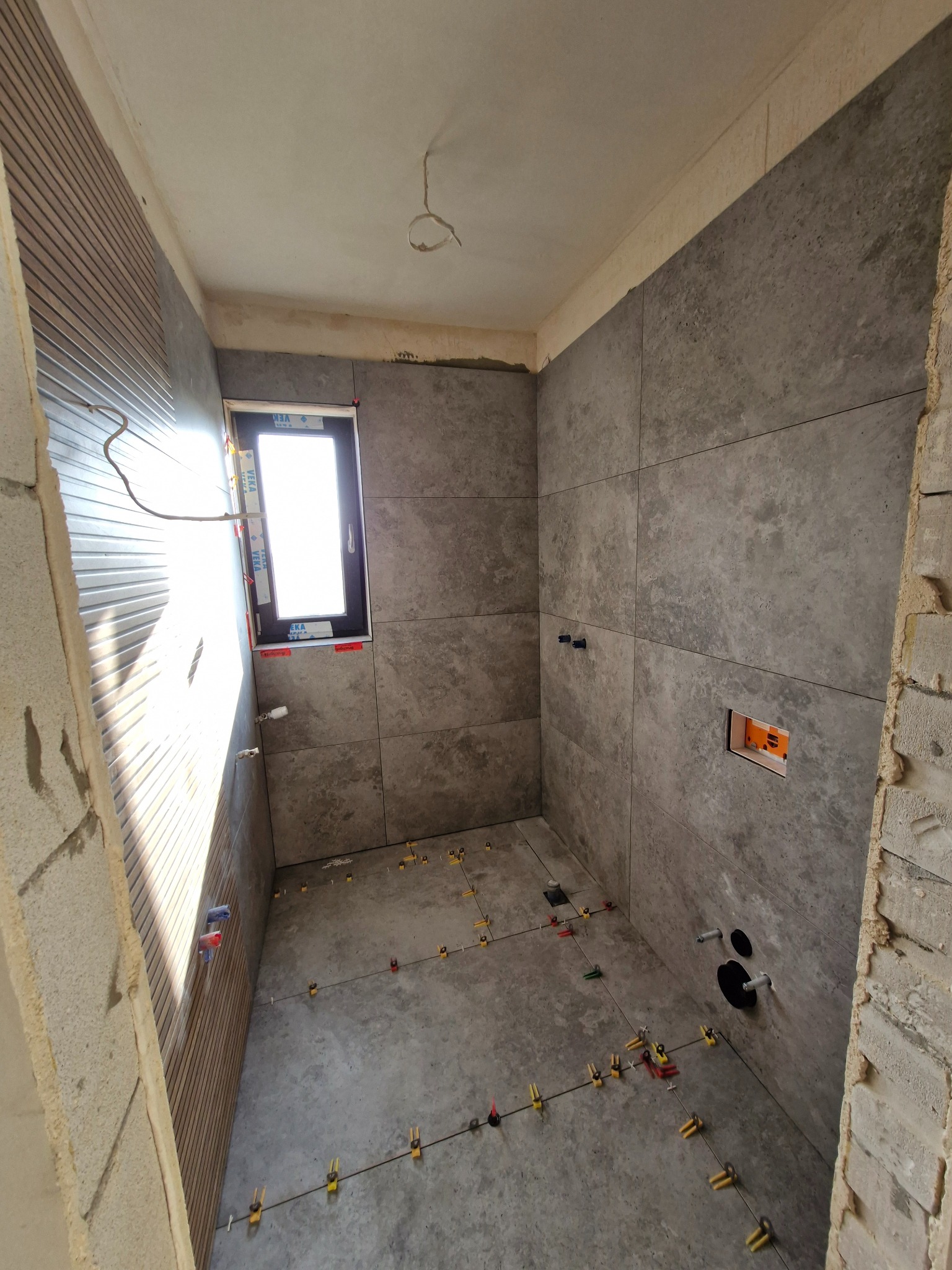 Łazienka w trakcie wykończenia: szare płytki na ścianach i podłodze, widoczne elementy instalacji, listwy wykończeniowe. Etap prac przed fugowaniem.