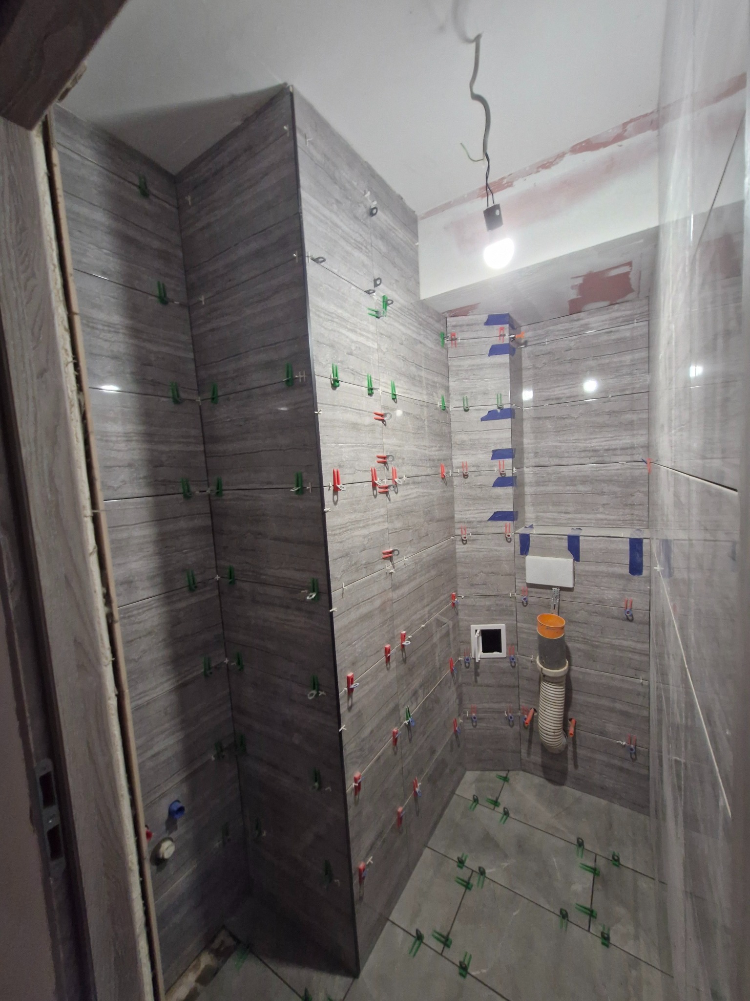 Łazienka w trakcie układania płytek imitujących drewno. Widoczne krzyżyki dystansowe. Częściowo zamontowana toaleta i odpływ. Oświetlenie sufitowe.