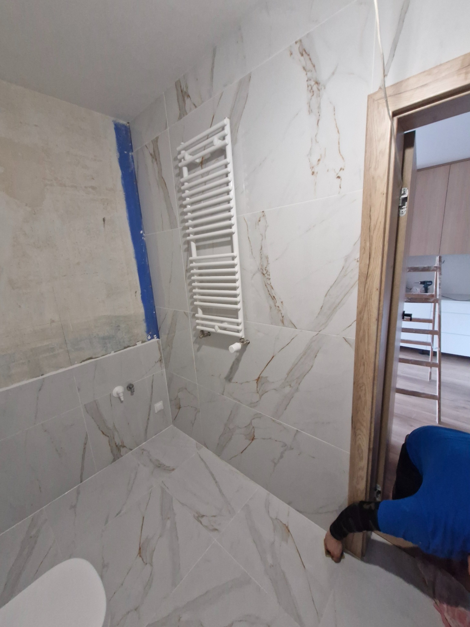 Łazienka w trakcie wykończenia: marmurowe płytki, biały grzejnik drabinkowy, futryna drzwi. W tle drabina i częściowo widoczna osoba w niebieskiej bluzie.