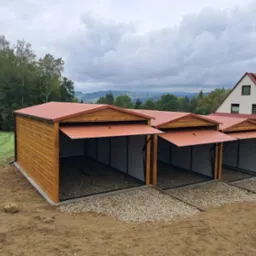 Trzy drewniane garaże blaszane z otwartymi bramami, ustawione jeden obok drugiego na żwirowym podłożu, z domem w tle i widokiem na zalesione wzgórza.