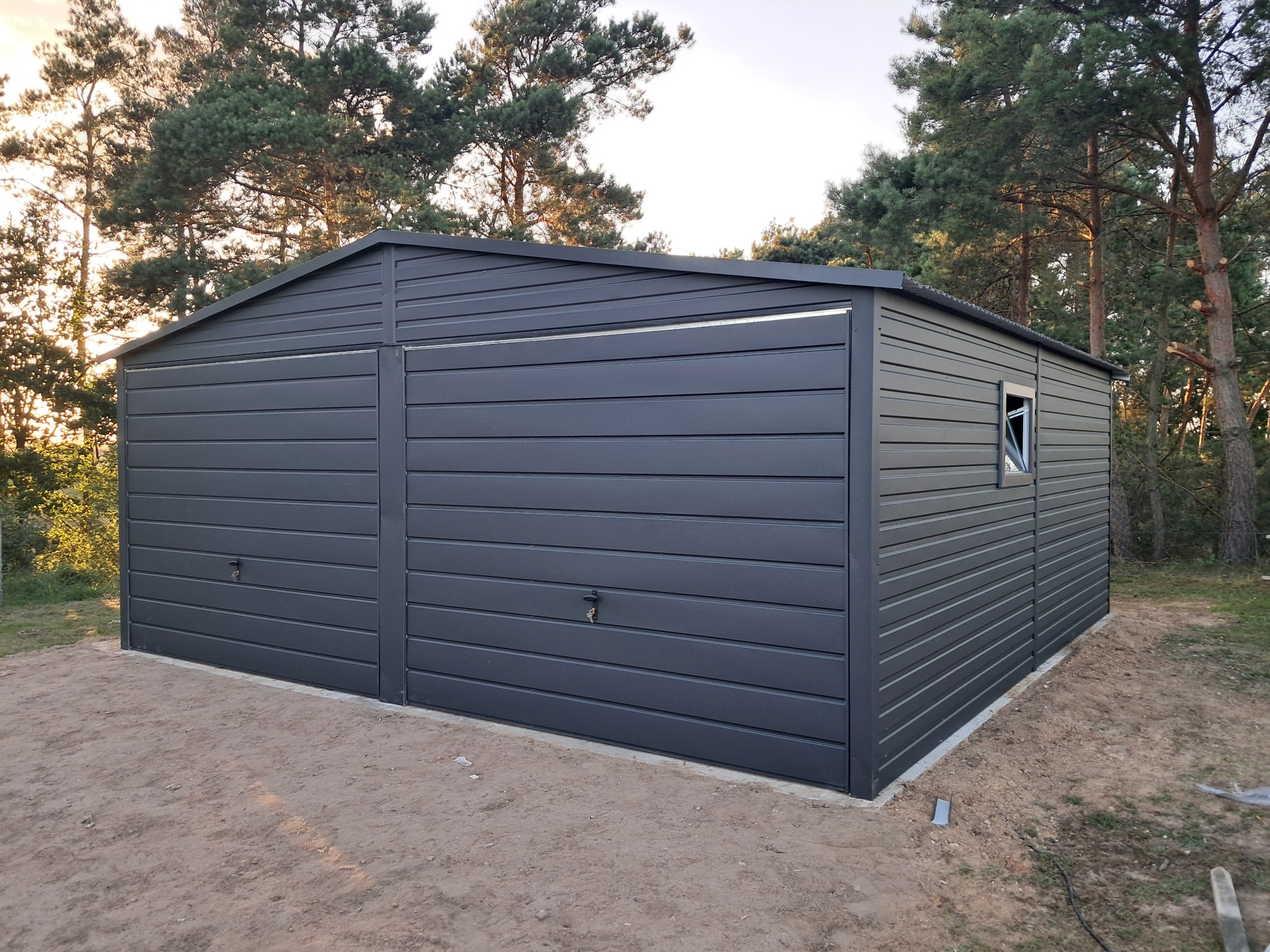 Nowoczesny, szary garaż blaszany z dwoma bramami segmentowymi, umieszczony na piaszczystym podłożu w otoczeniu drzew iglastych. Widoczne okno boczne.
