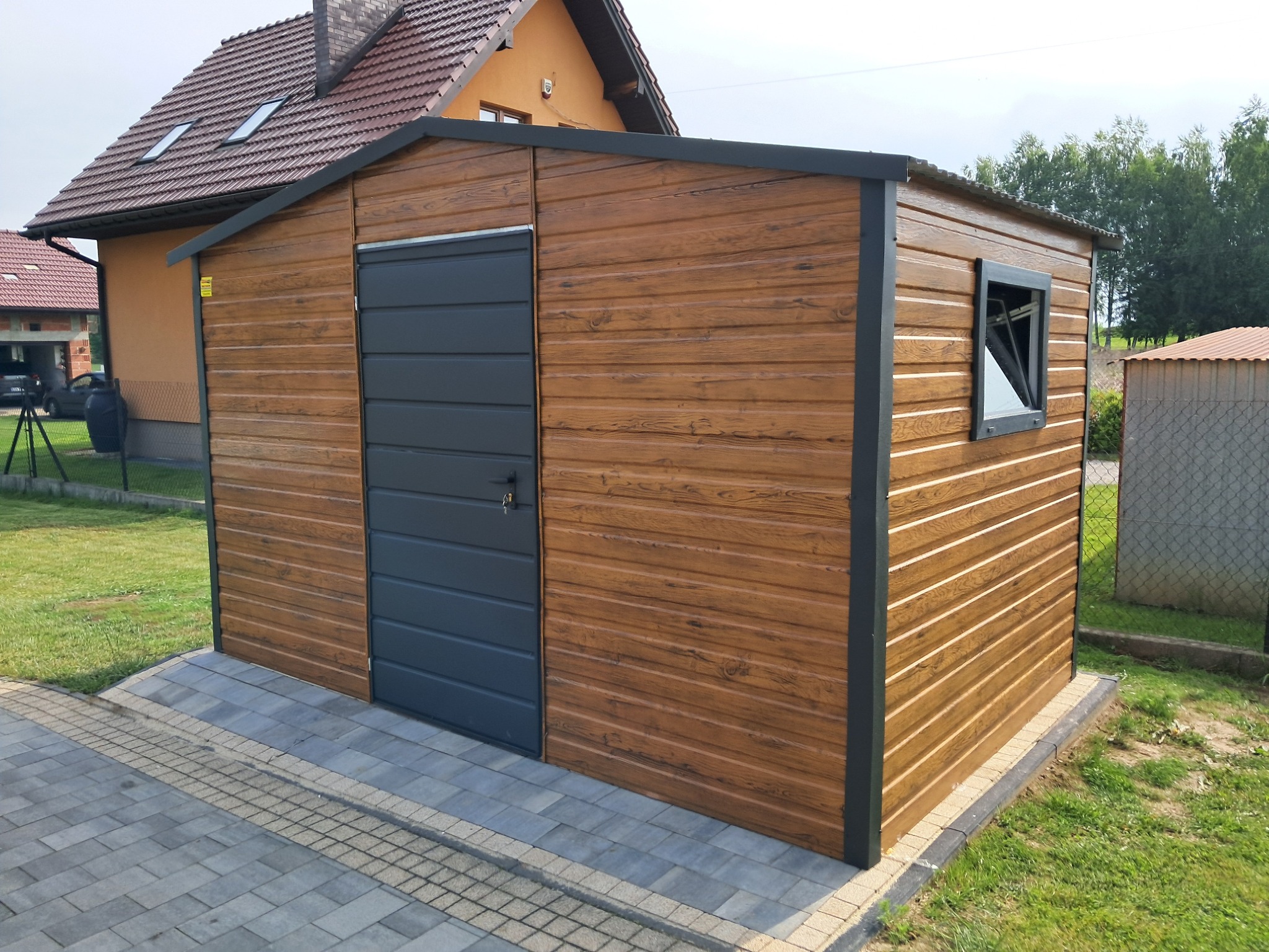 Gotowy garaż blaszany z drewnopodobną elewacją i szarą bramą, umieszczony na kostce brukowej obok domu jednorodzinnego. Widok na ścianę z oknem.