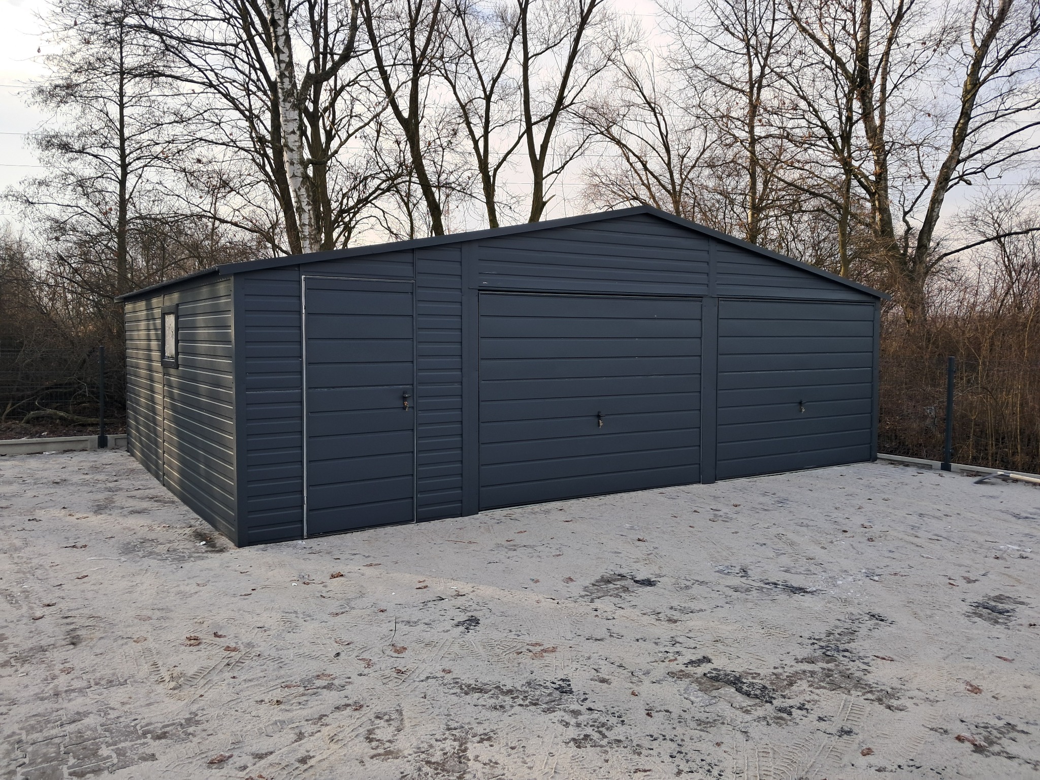 Nowoczesny garaż blaszany w grafitowym kolorze, z trzema segmentowymi bramami i oknem, na betonowej wylewce, otoczony drzewami bez liści.