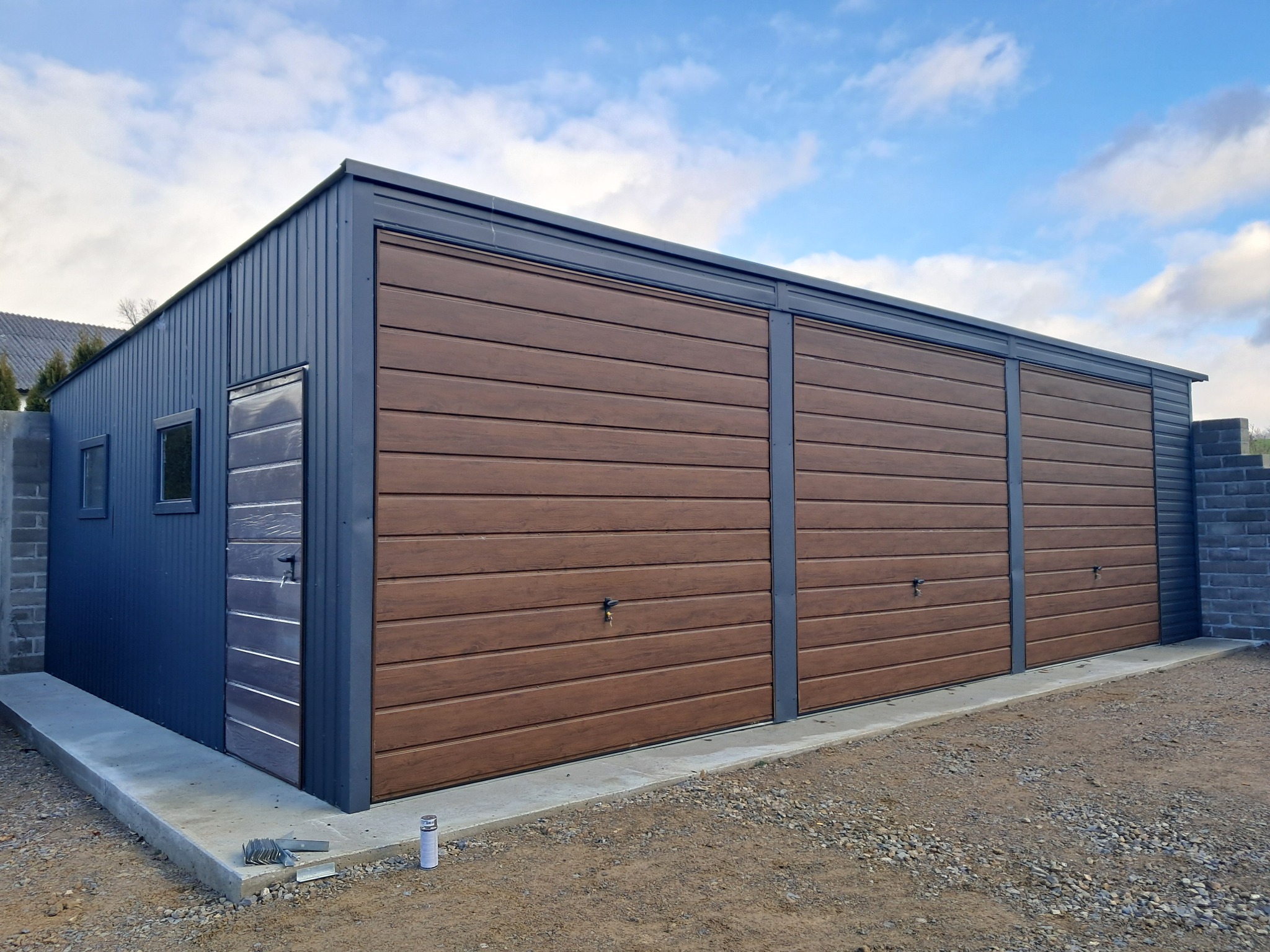 Nowoczesny garaż blaszany z dwiema bramami segmentowymi o strukturze drewna, szara blacha trapezowa, widoczne narzędzia i puszka farby na żwirowym podłożu.