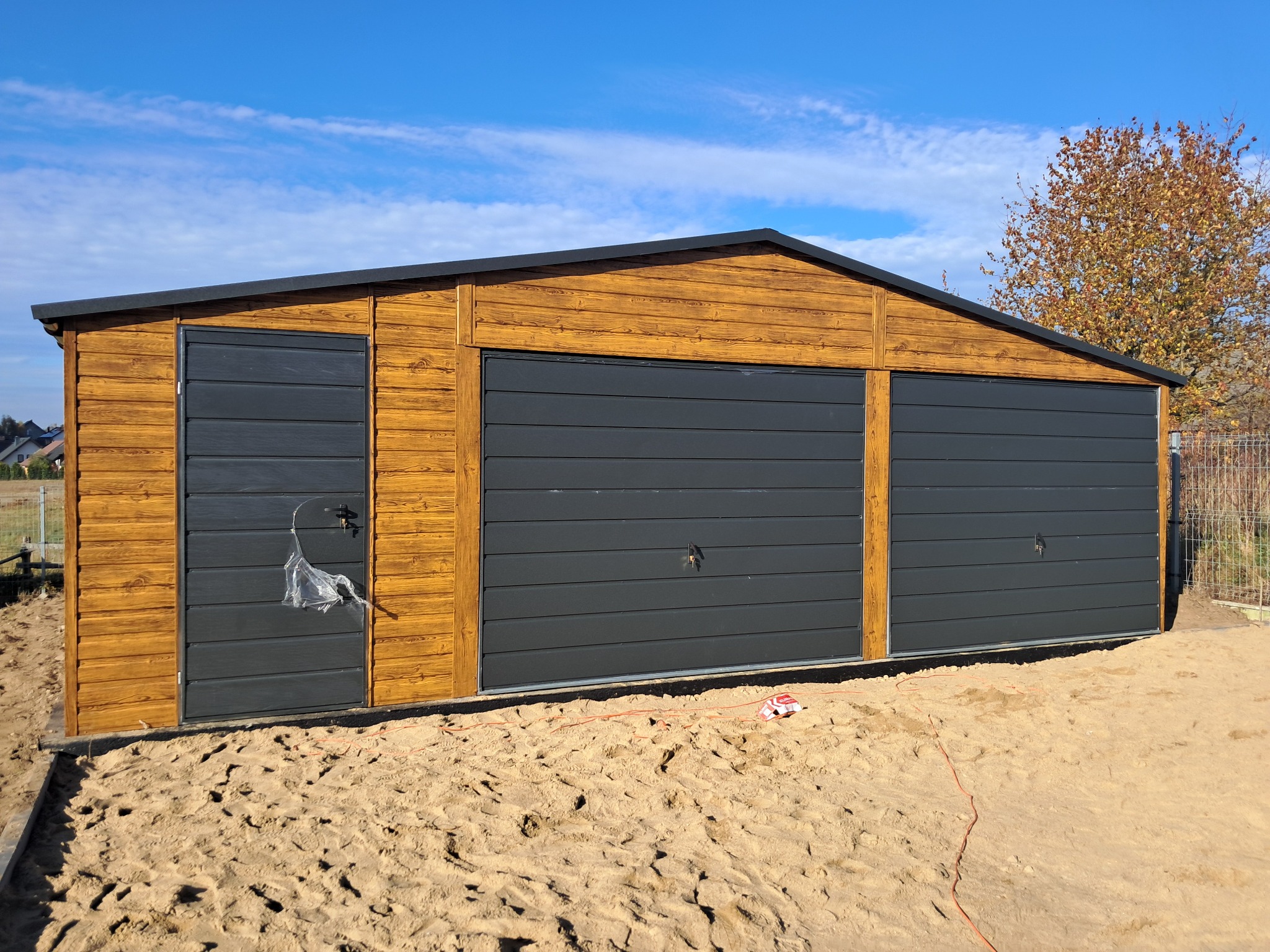 Garaż blaszany z trzema bramami segmentowymi w kolorze grafitowym, wykończony drewnianą boazerią. Widoczne ślady montażu w piasku.