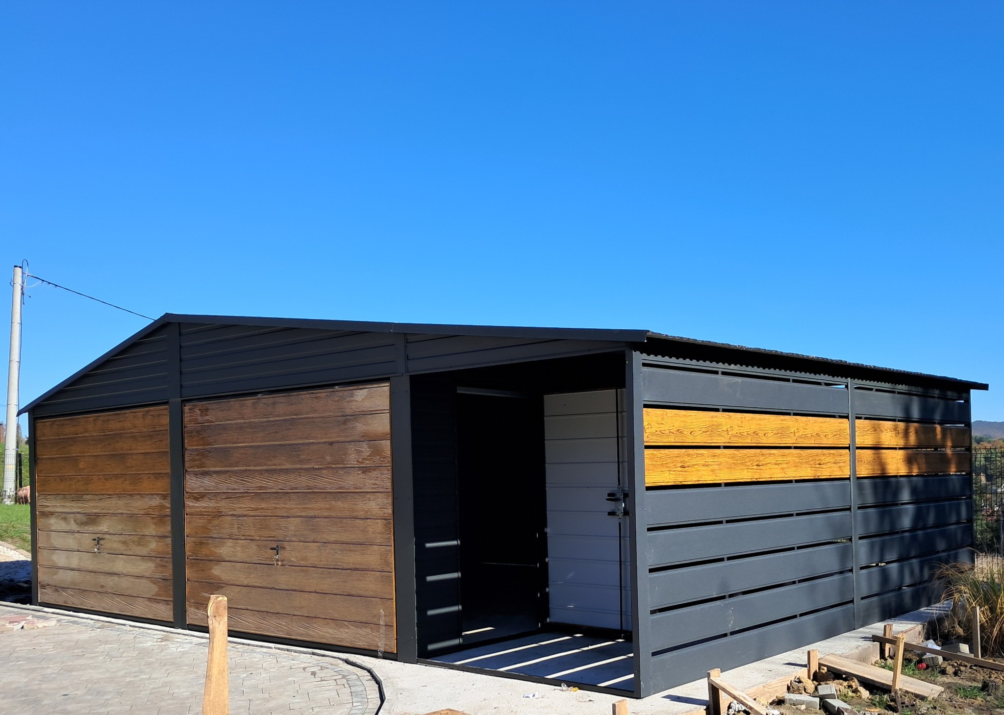 Nowoczesny garaż blaszany z drewnopodobnymi bramami przesuwnymi i elementami dekoracyjnymi w kolorze drewna na tle błękitnego nieba.