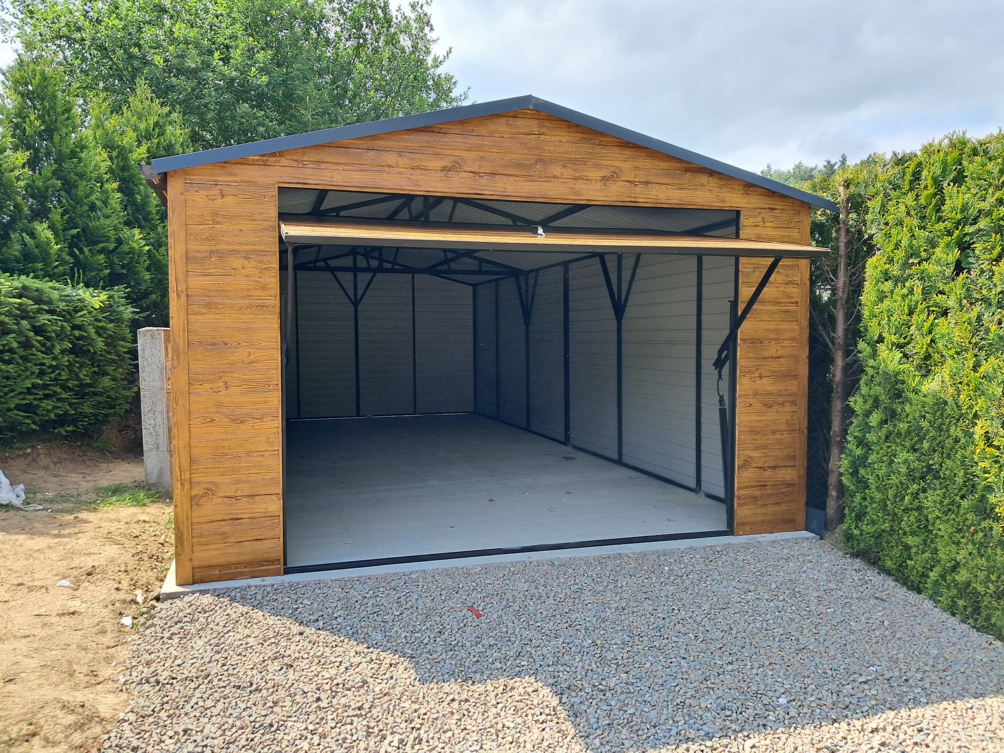 Otwarty garaż blaszany z imitacją drewna, widok z zewnątrz na żwirowe podłoże i zielone krzewy. Konstrukcja z metalu, brama uchylna w połowie otwarta.