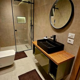 EKSBUD SPÓŁKA Z OGRANICZONĄ ODPOWIEDZIALNOŚCIĄ - Nowoczesna łazienka z kabiną prysznicową typu walk-in, czarną umywalką nablatową i okrągłym lustrem. Beżowe płytki, oświetlenie LED i brązowe dywaniki.