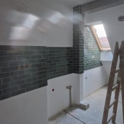 EKSBUD SPÓŁKA Z OGRANICZONĄ ODPOWIEDZIALNOŚCIĄ - Wnętrze w trakcie wykańczania, z ułożonymi ciemnozielonymi płytkami na ścianach do połowy wysokości i drewnianą drabiną opartą o ścianę przy oknie dachowym.