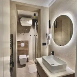 Jasna łazienka po remoncie: beżowe płytki, okrągłe lustro z podświetleniem, wisząca toaleta, kabina prysznicowa i czarne akcenty oświetleniowe.