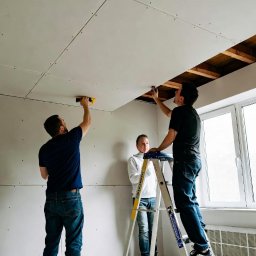 EKSBUD SPÓŁKA Z OGRANICZONĄ ODPOWIEDZIALNOŚCIĄ - Montaż płyt karton-gips na suficie przez dwóch mężczyzn, trzeci przygląda się z drabiny. Widoczne drewniane belki i okno w tle. Praca w jasnym pomieszczeniu.