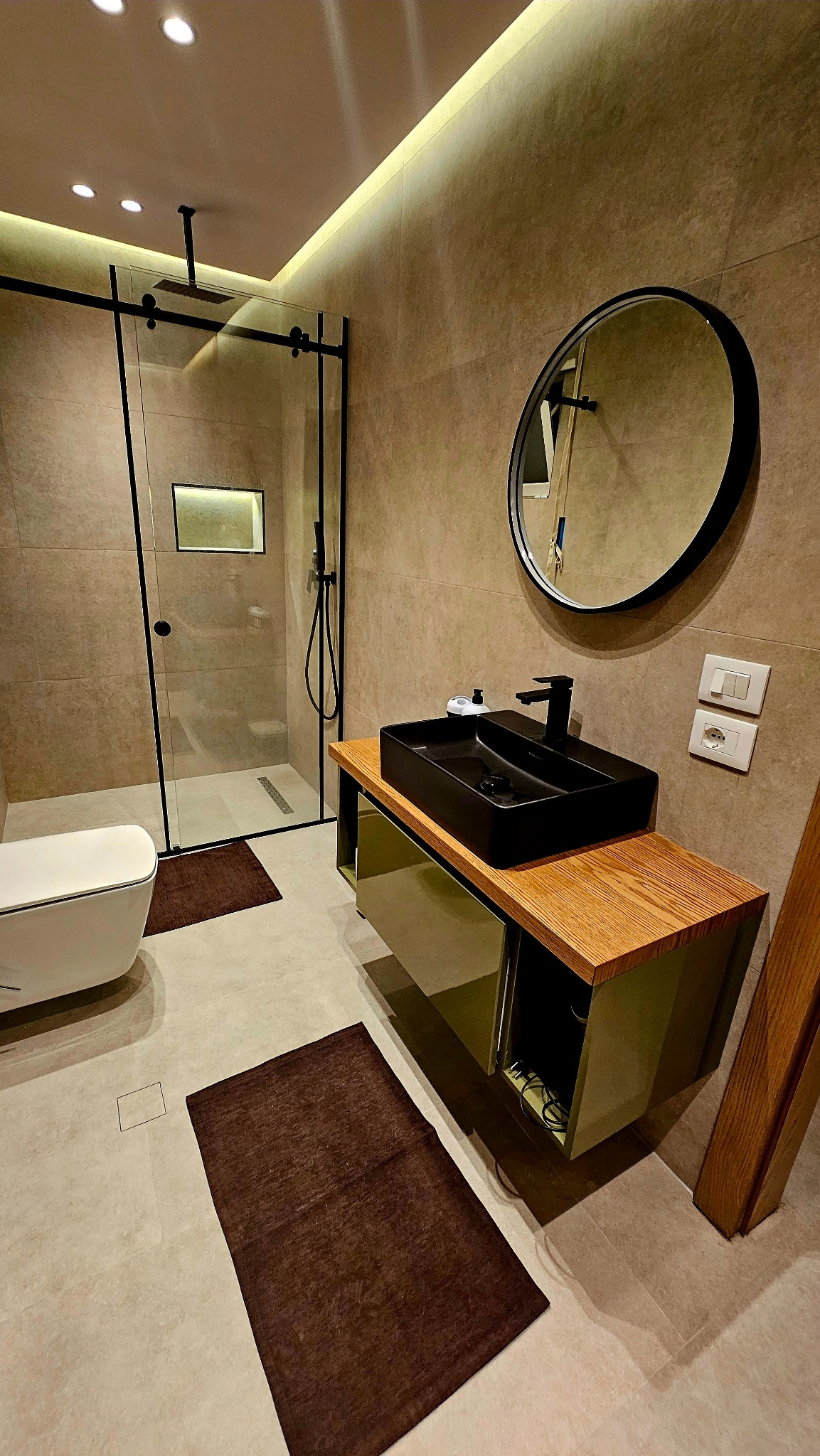 Nowoczesna łazienka z kabiną prysznicową typu walk-in, czarną umywalką nablatową i okrągłym lustrem. Beżowe płytki, oświetlenie LED i brązowe dywaniki.