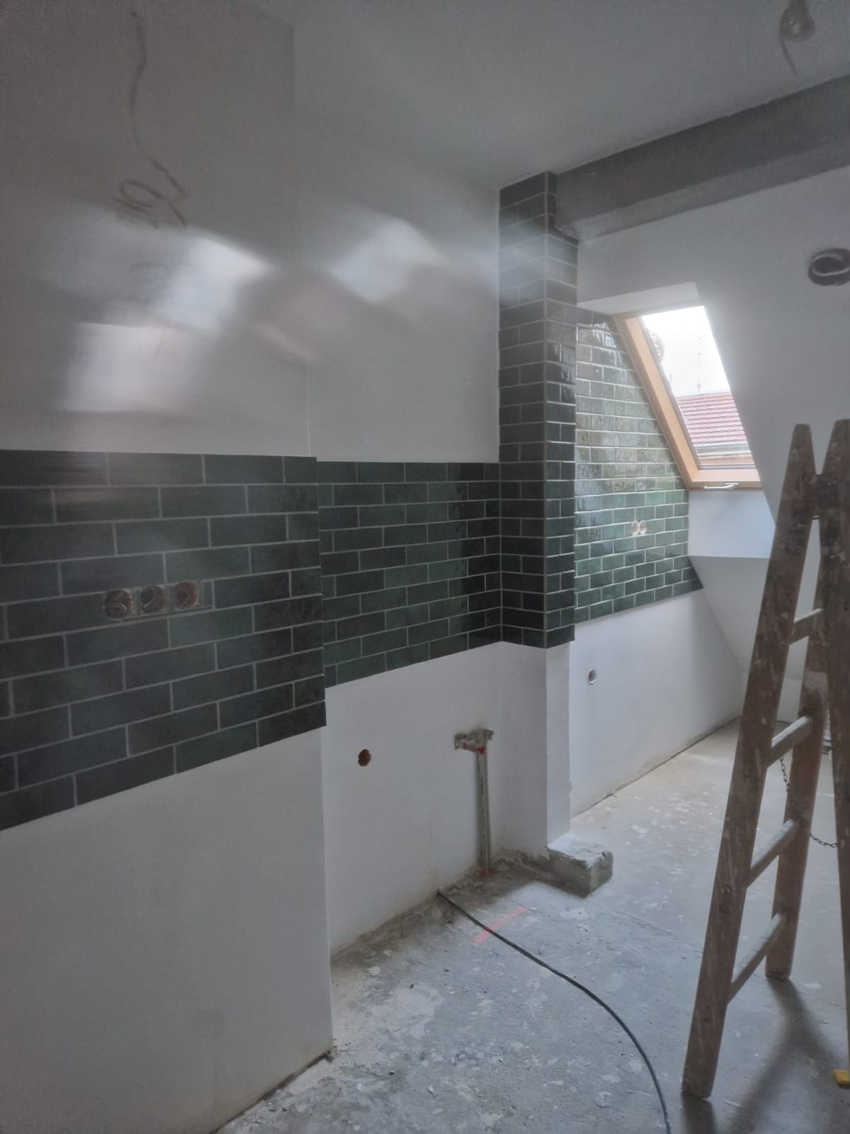 Wnętrze w trakcie wykańczania, z ułożonymi ciemnozielonymi płytkami na ścianach do połowy wysokości i drewnianą drabiną opartą o ścianę przy oknie dachowym.