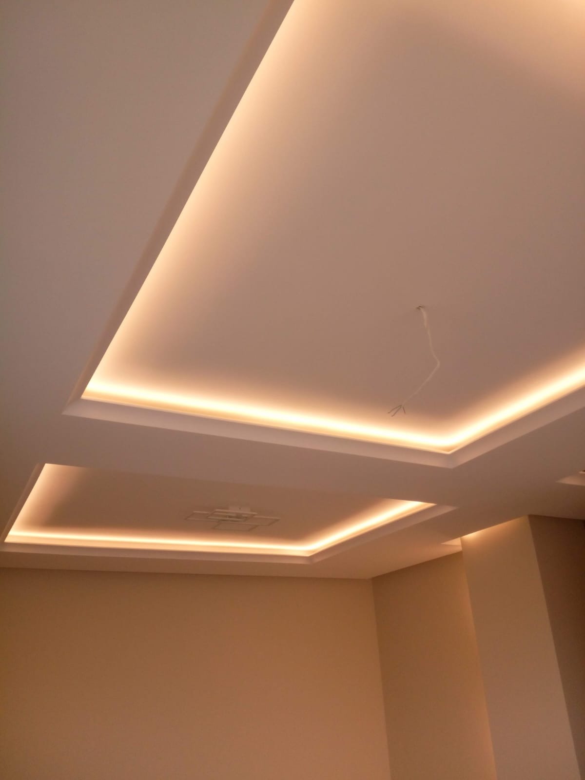Dwupoziomowy sufit podwieszany z oświetleniem LED w ciepłej barwie, widok z dołu. Kremowe ściany i wystający kabel elektryczny.