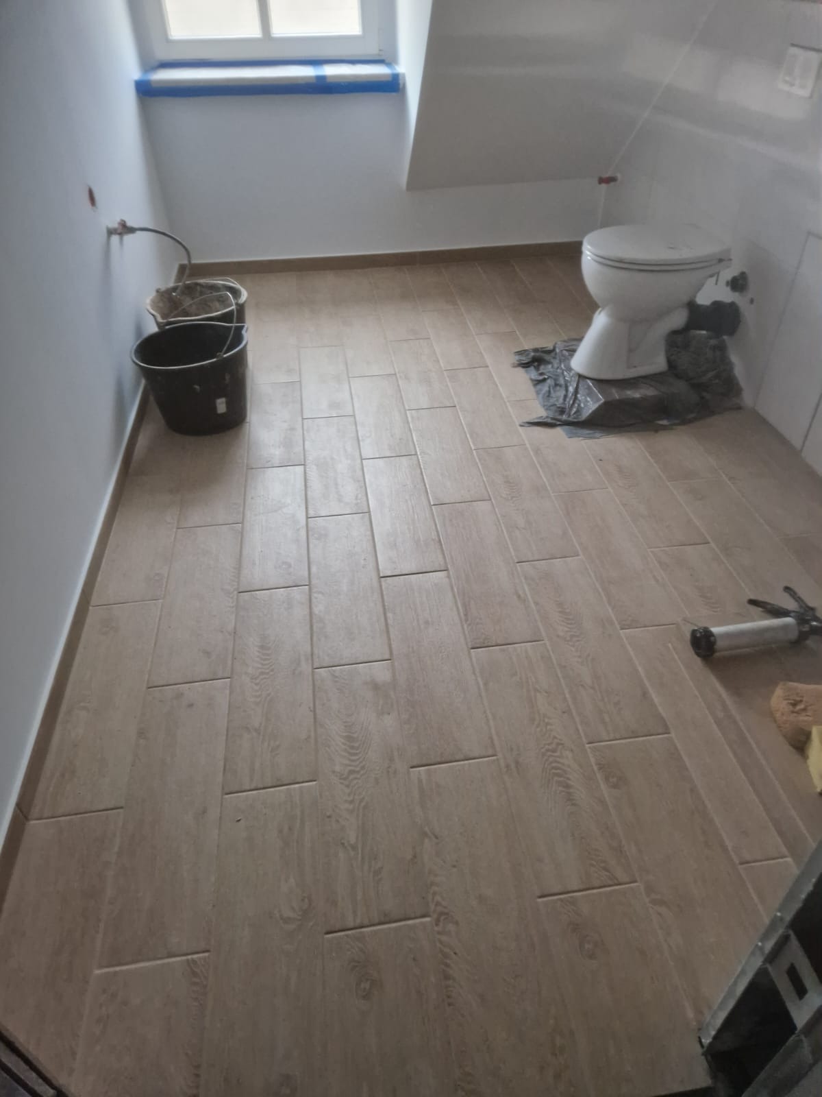 Ułożona podłoga z płytek imitujących drewno w łazience podczas remontu. Widoczne narzędzia i wiadro, przygotowany sedes. Okno zabezpieczone folią.