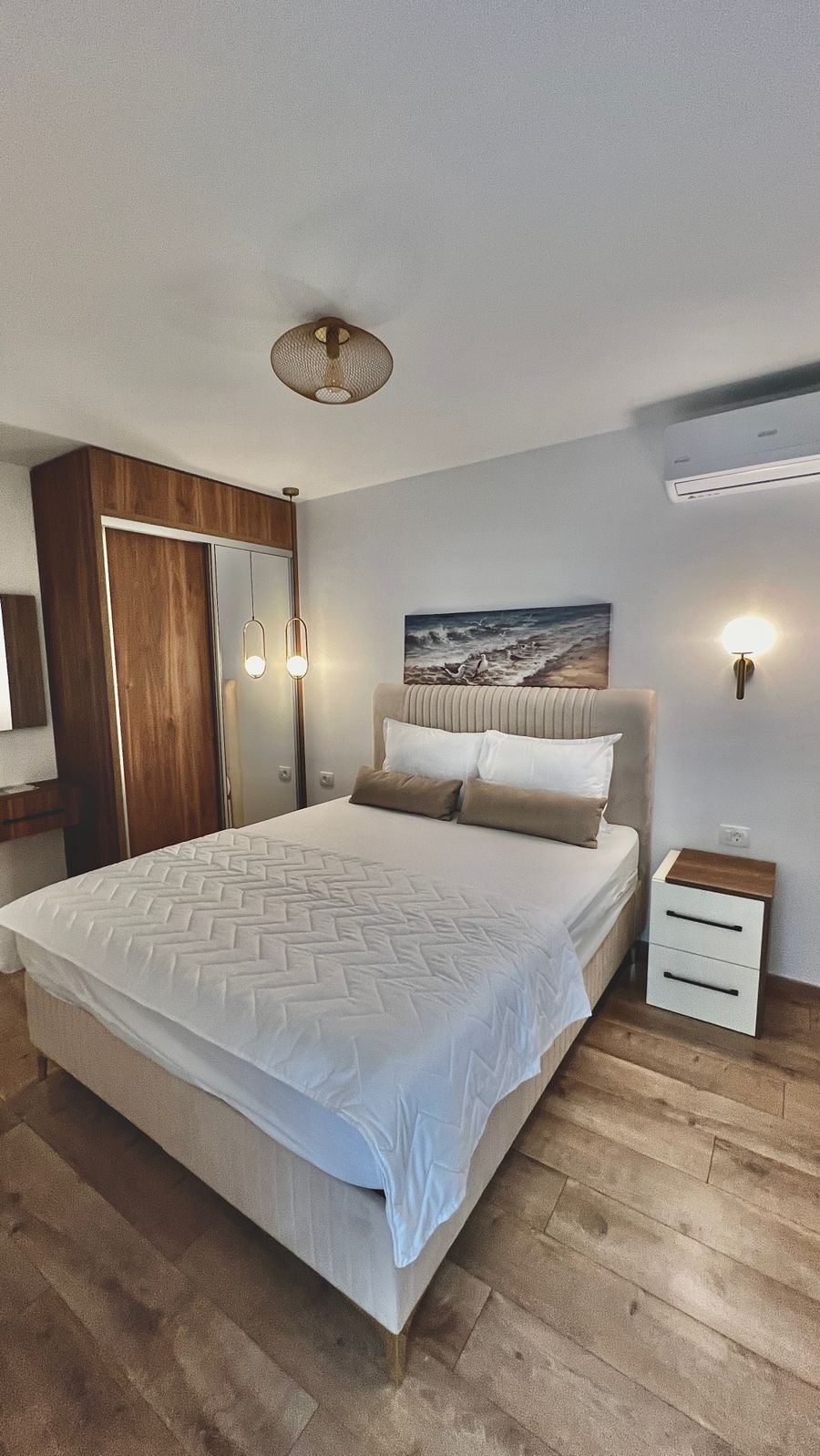 Jasna sypialnia z drewnianą podłogą i szafą wnękową. Nad łóżkiem obraz z motywem morza. Na ścianie kinkiet i klimatyzator.  Wnętrze po remoncie.