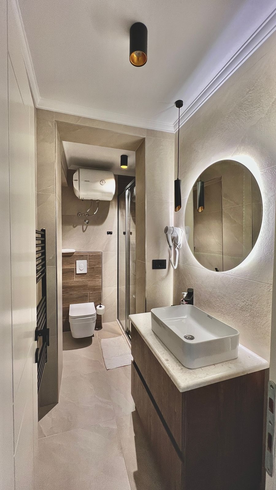 Jasna łazienka po remoncie: beżowe płytki, okrągłe lustro z podświetleniem, wisząca toaleta, kabina prysznicowa i czarne akcenty oświetleniowe.