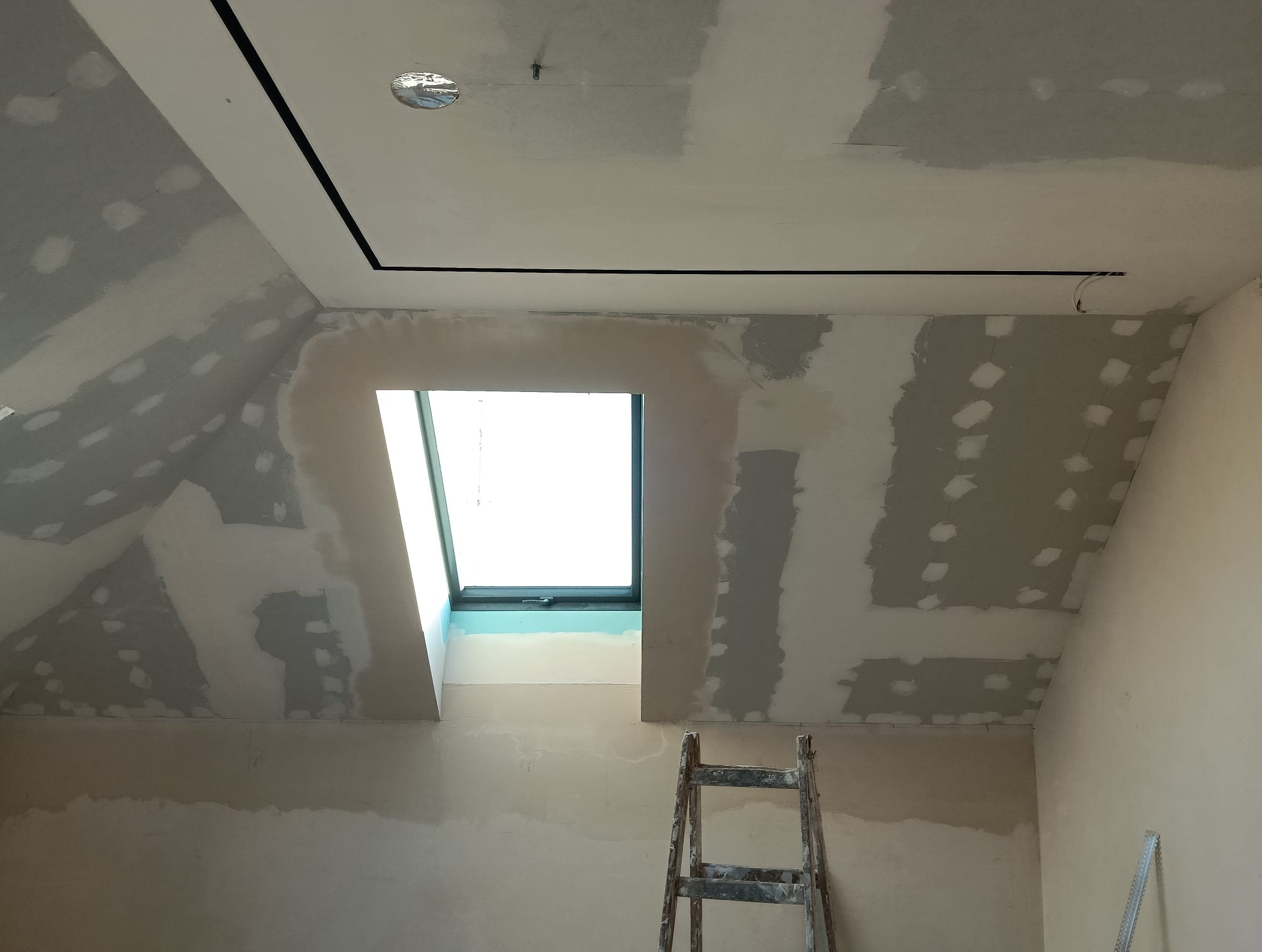 Poddasze z oknem dachowym w trakcie prac wykończeniowych. Widoczne szpachlowane ściany, sufit z czarną listwą i drabina. Etap przygotowania do malowania.