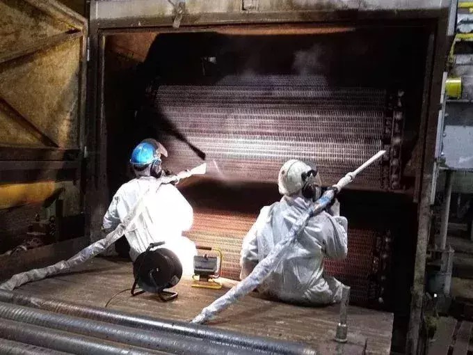 Dwóch pracowników w kombinezonach czyszczących przemysłowy wymiennik ciepła wewnątrz metalowej komory, używając specjalistycznych pistoletów natryskowych.