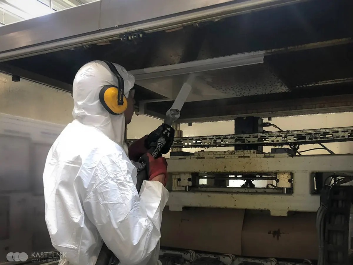Pracownik w kombinezonie ochronnym czyści przemysłową konstrukcję za pomocą specjalistycznego sprzętu pod ciśnieniem. Ochrona słuchu i rękawice zapewniają bezpieczeństwo.