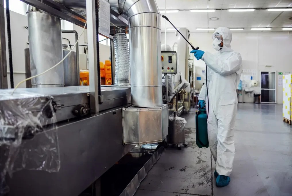 Dezynfekcja przemysłowa: pracownik w kombinezonie ochronnym, z maską i rękawicami, spryskuje metalową maszynę w hali produkcyjnej. Widoczne elementy wentylacji.