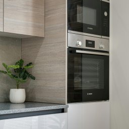 Fidelis Group Bartłomiej Wierzbicki - Nowoczesna zabudowa kuchenna z drewnopodobnymi panelami, sprzętem Bosch i rośliną w białej doniczce. Minimalistyczny design, jasne kolory.
