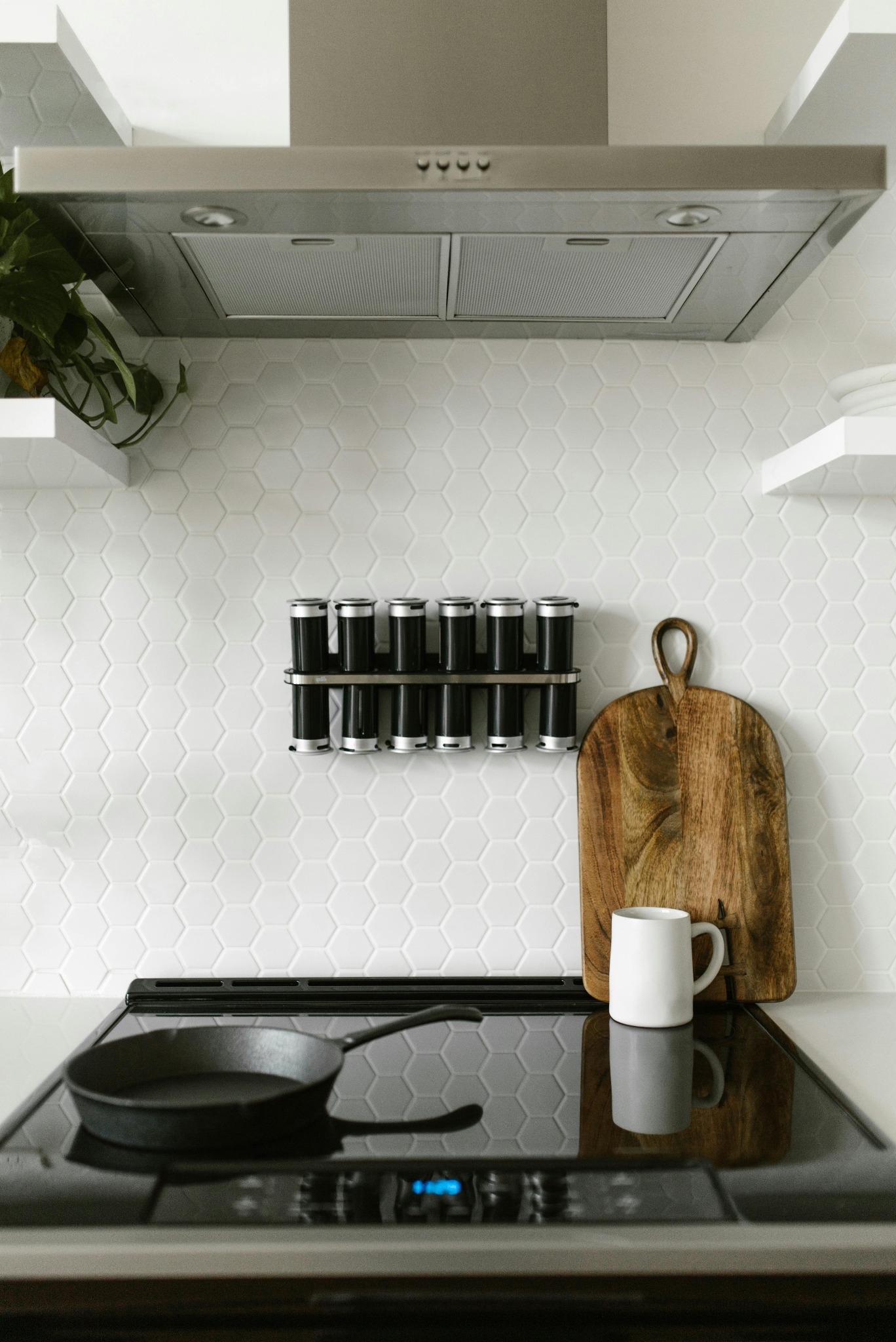Uporządkowana kuchnia: czarna patelnia na płycie, drewniana deska, kubek i przyprawy na tle białych heksagonalnych płytek i stalowego okapu.