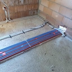 4Life KRYSTIAN RADOSZ - Instalacja ogrzewania podłogowego: Rury grzewcze w kolorach czerwonym i niebieskim ułożone na podłodze w surowym wnętrzu, przed wylaniem posadzki. Widoczne przewody elektryczne.