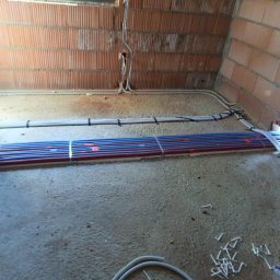 4Life KRYSTIAN RADOSZ - Surowy stan: rury ogrzewania podłogowego (czerwono-niebieskie) i kable elektryczne ułożone na podłodze w pomieszczeniu z ceglanymi ścianami przed wylaniem wylewki.