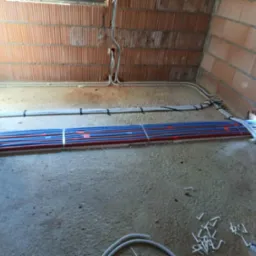 Surowy stan: rury ogrzewania podłogowego (czerwono-niebieskie) i kable elektryczne ułożone na podłodze w pomieszczeniu z ceglanymi ścianami przed wylaniem wylewki.