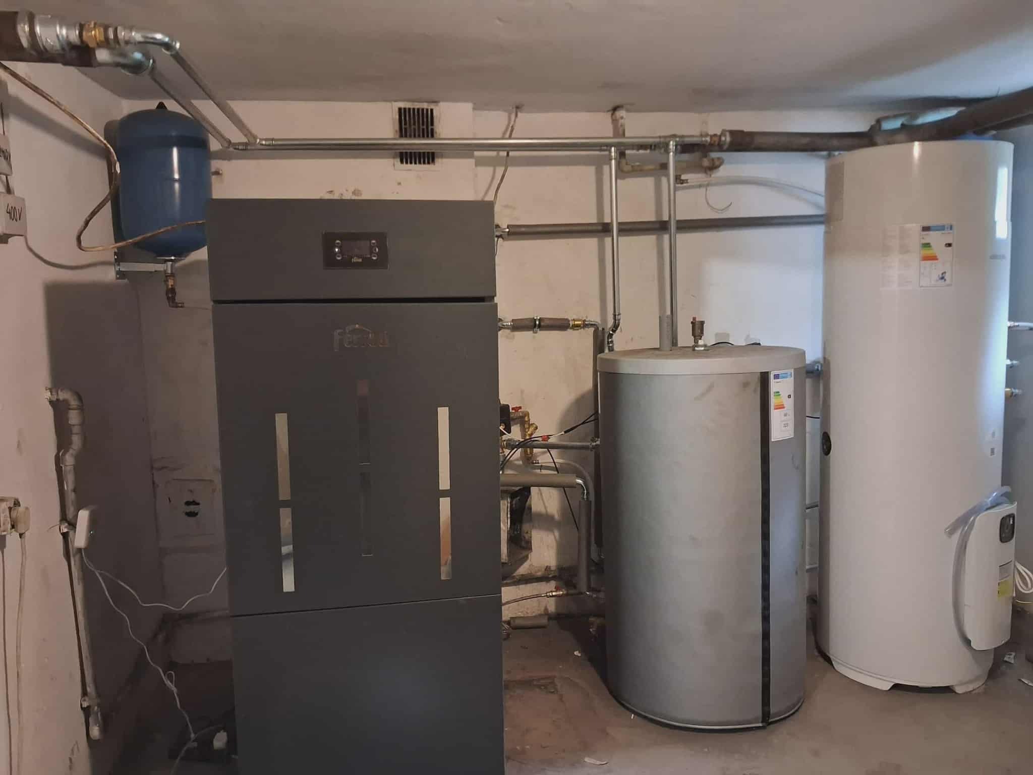 Zestaw urządzeń grzewczych w piwnicy: piec z wyświetlaczem, zbiornik buforowy, bojler. Widoczne rury i instalacje. Oświetlenie piwniczne, surowy beton na podłodze.