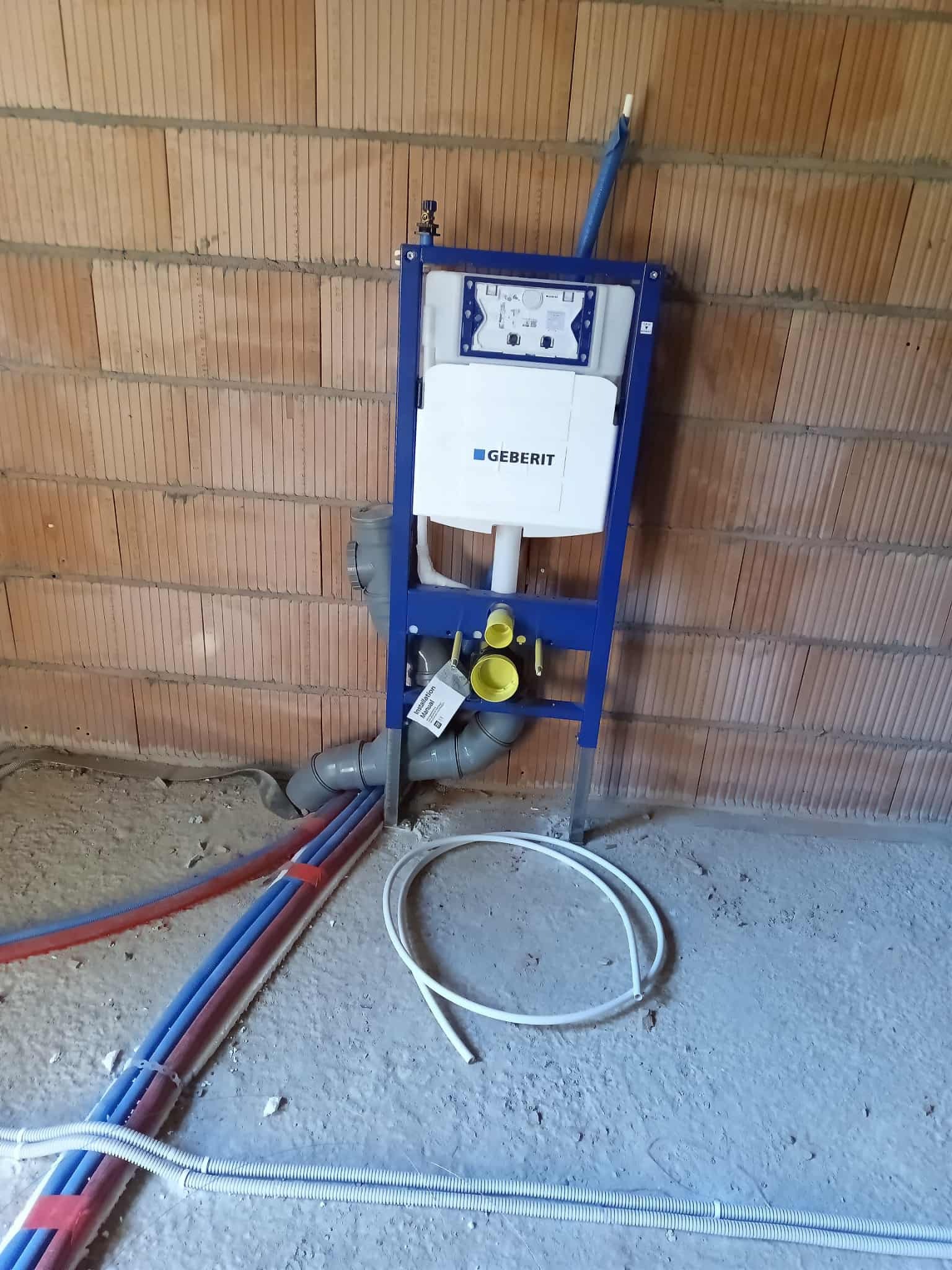 Stelaż podtynkowy Geberit zamontowany na tle ściany z czerwonej cegły. Widoczne rury doprowadzające wodę i odpływ kanalizacyjny. Podłoże betonowe.