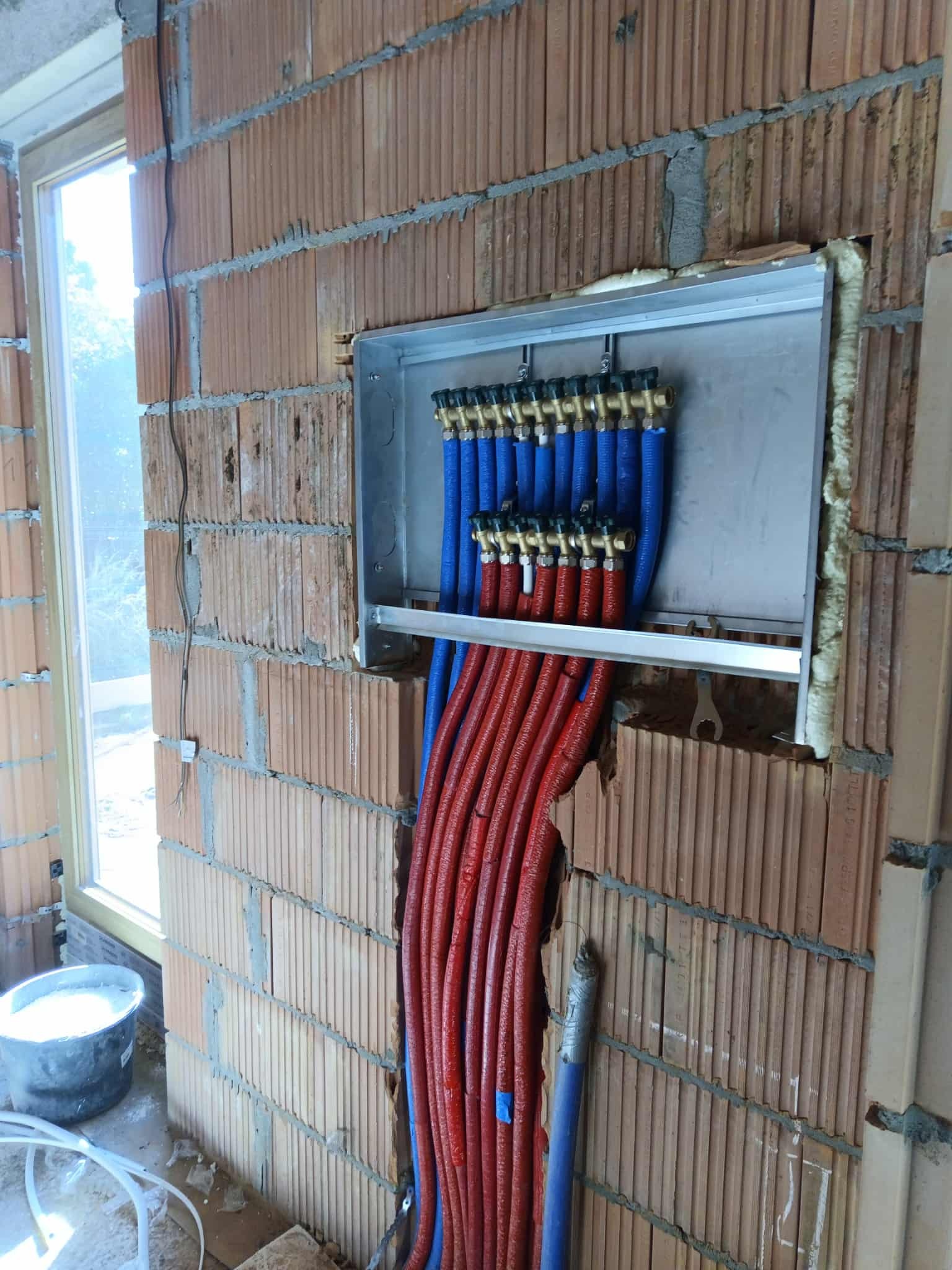 Rozdzielacz ogrzewania podłogowego zamontowany w ścianie z cegły, z widocznymi rurami w kolorze czerwonym i niebieskim, prowadzącymi do instalacji.