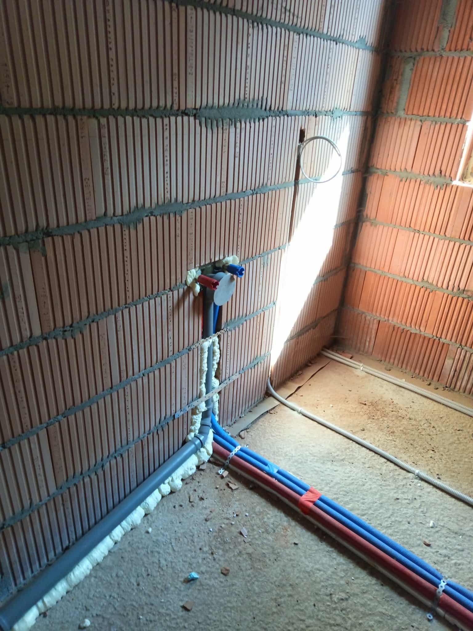 Instalacja wodna w surowym wnętrzu: czerwono-niebieskie rury prowadzone wzdłuż ściany z cegły, zabezpieczone pianką montażową. Widoczne przyłącza i fragment podłogi.