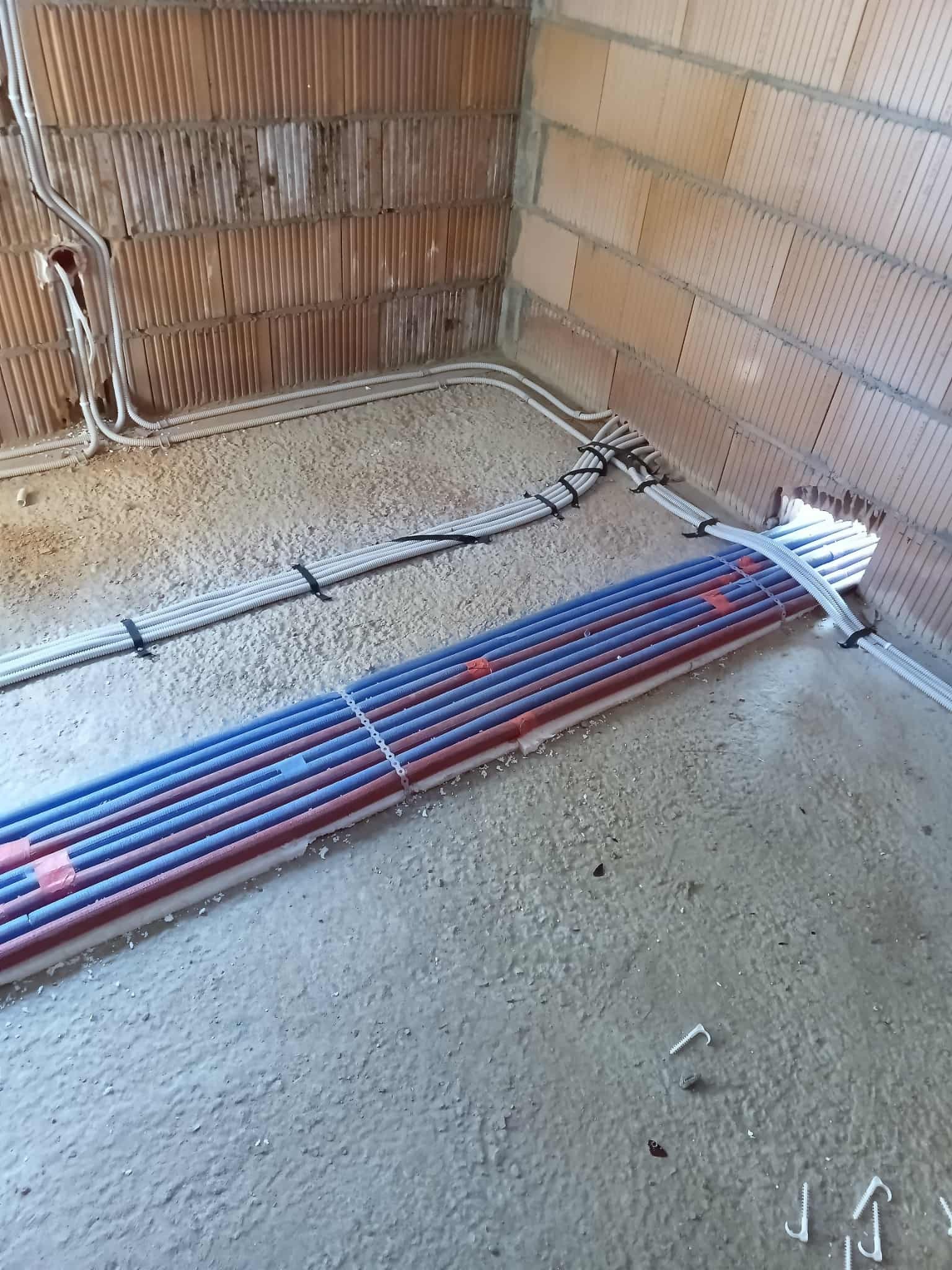 Instalacja ogrzewania podłogowego: Rury grzewcze w kolorach czerwonym i niebieskim ułożone na podłodze w surowym wnętrzu, przed wylaniem posadzki. Widoczne przewody elektryczne.