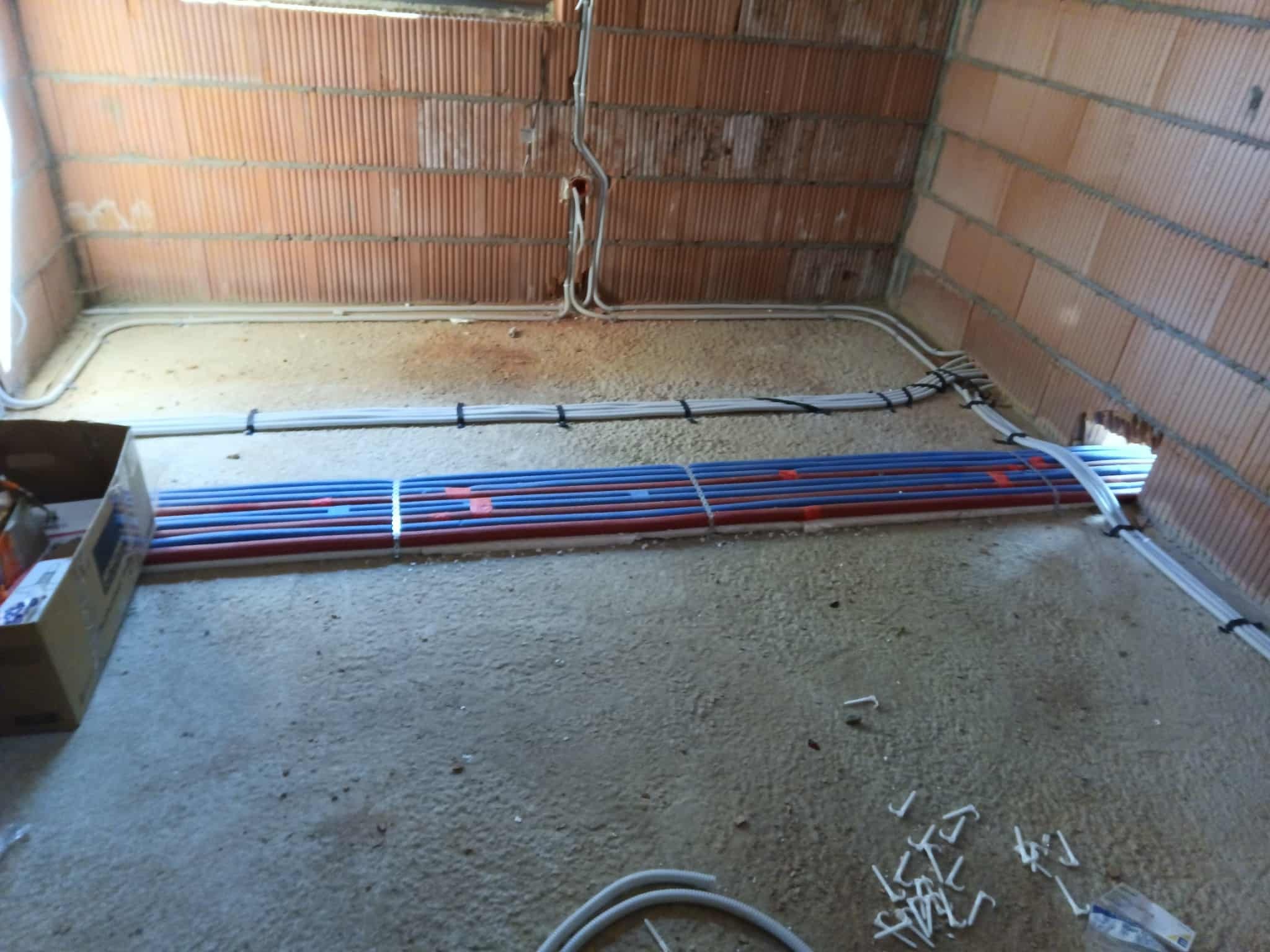 Surowy stan: rury ogrzewania podłogowego (czerwono-niebieskie) i kable elektryczne ułożone na podłodze w pomieszczeniu z ceglanymi ścianami przed wylaniem wylewki.