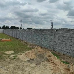 Ogrodzenia Kamil - Długi, szary, betonowy płot z dekoracyjnym wzorem kamienia i łukowatym wykończeniem, oddzielający posesję. Na pierwszym planie hałda piasku.