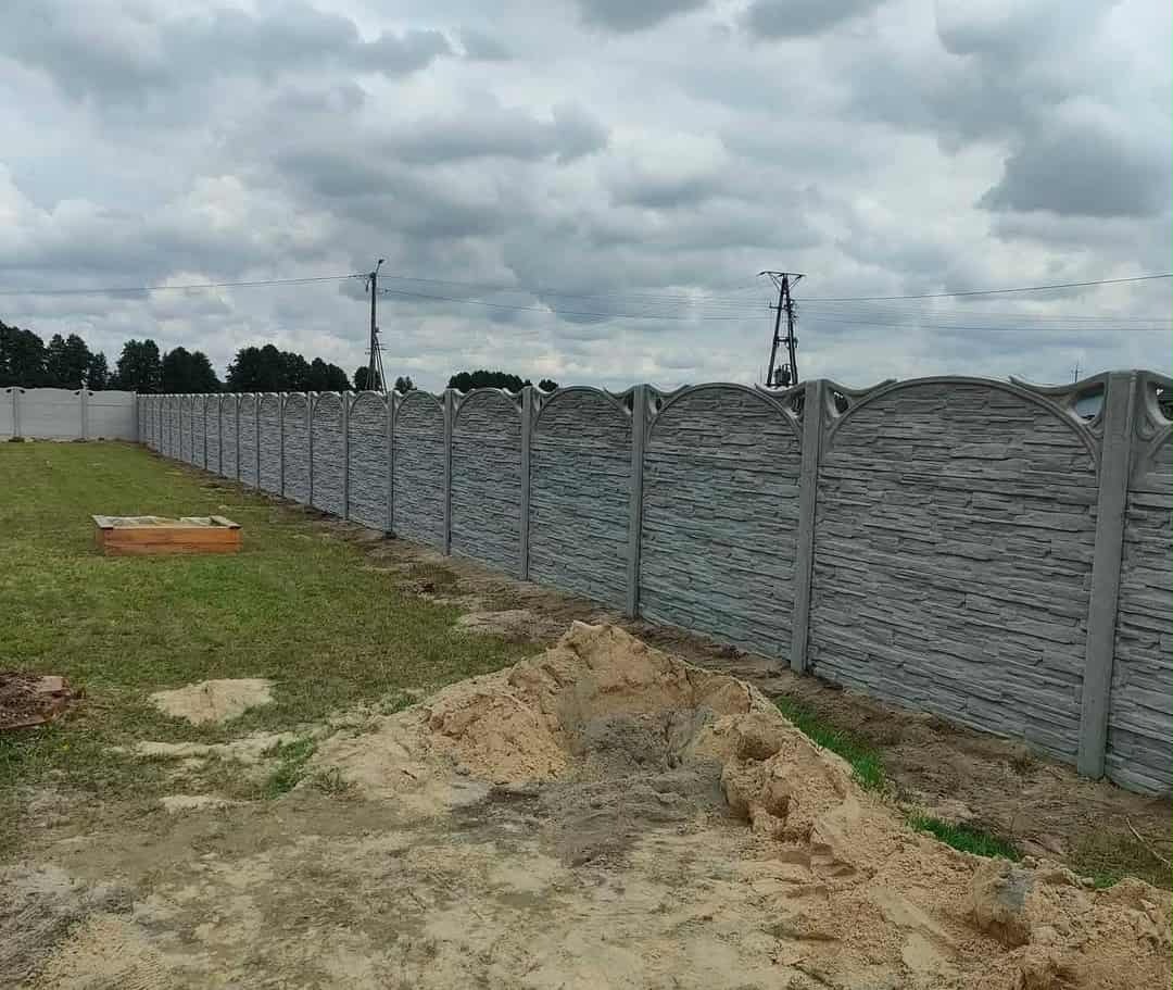 Długi, szary, betonowy płot z dekoracyjnym wzorem kamienia i łukowatym wykończeniem, oddzielający posesję. Na pierwszym planie hałda piasku.