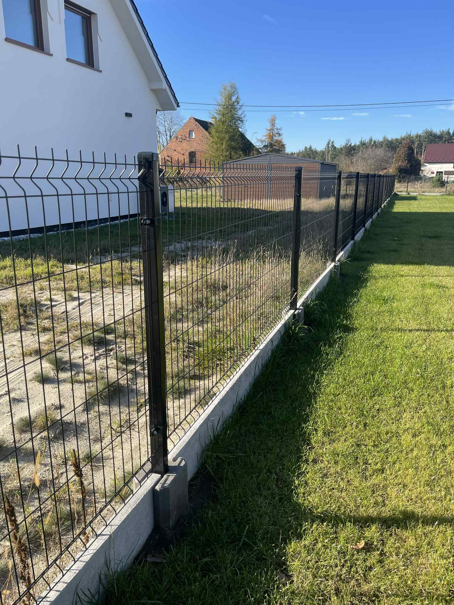 Długi, czarny panel ogrodzeniowy na betonowej podmurówce, oddzielający trawnik od terenu z piaskiem i chwastami. W tle dom z białymi ścianami i czerwonym dachem.