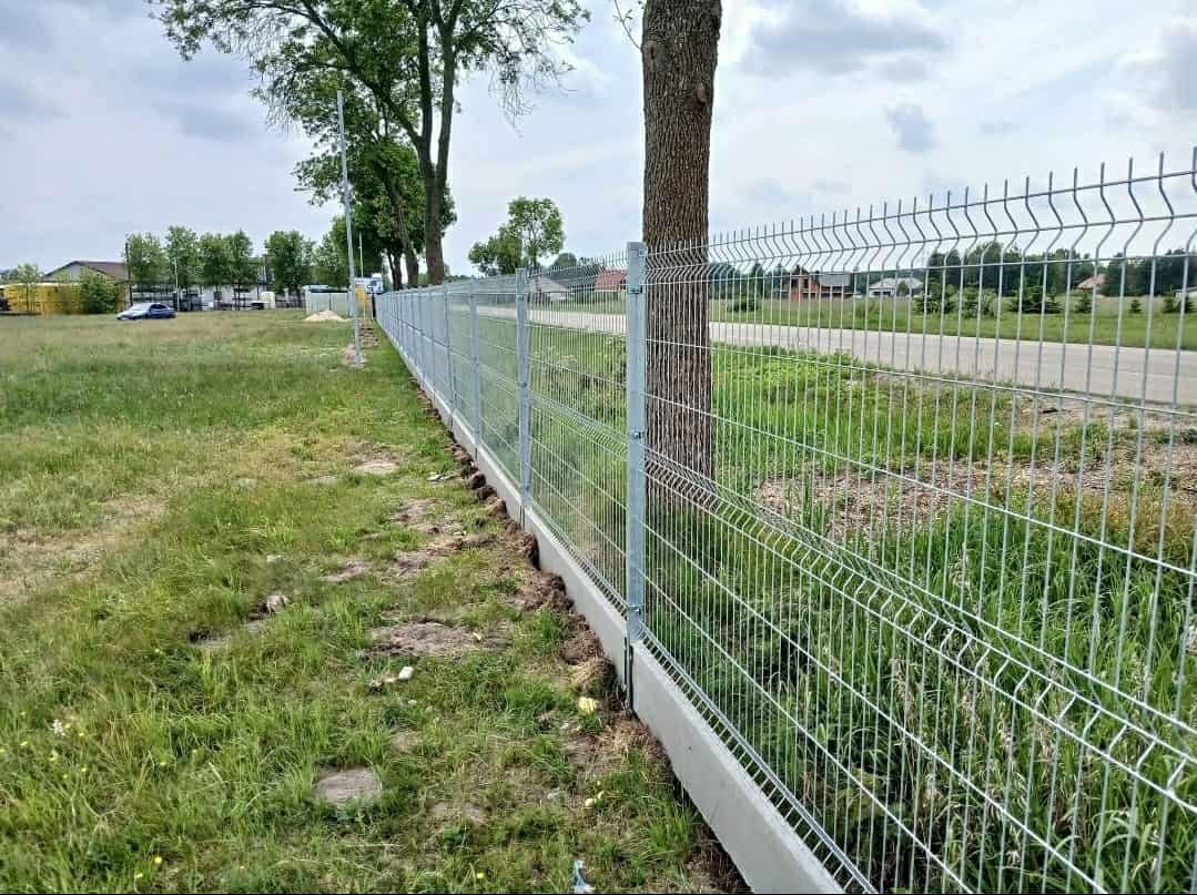 Długi, srebrny panel ogrodzeniowy z betonową podmurówką, biegnący wzdłuż trawnika i drzewa. Widok na drogę i zabudowania w tle, pochmurne niebo.