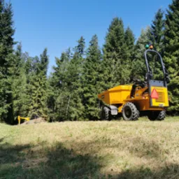 Żółta wywrotka Thwaites 3 tonne na tle lasu iglastego i błękitnego nieba. W tle widoczna koparka. Prace ziemne w toku, słoneczny dzień.